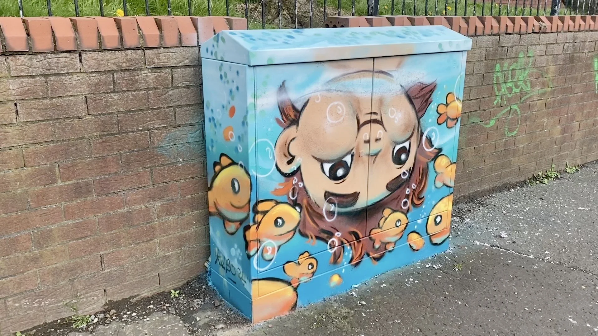 Colorful graffiti mural on a utility box depicting a smiling cartoon girl with brown hair surrounded by orange fish, set against a blue background with bubbles and flower accents, next to a brick wall.