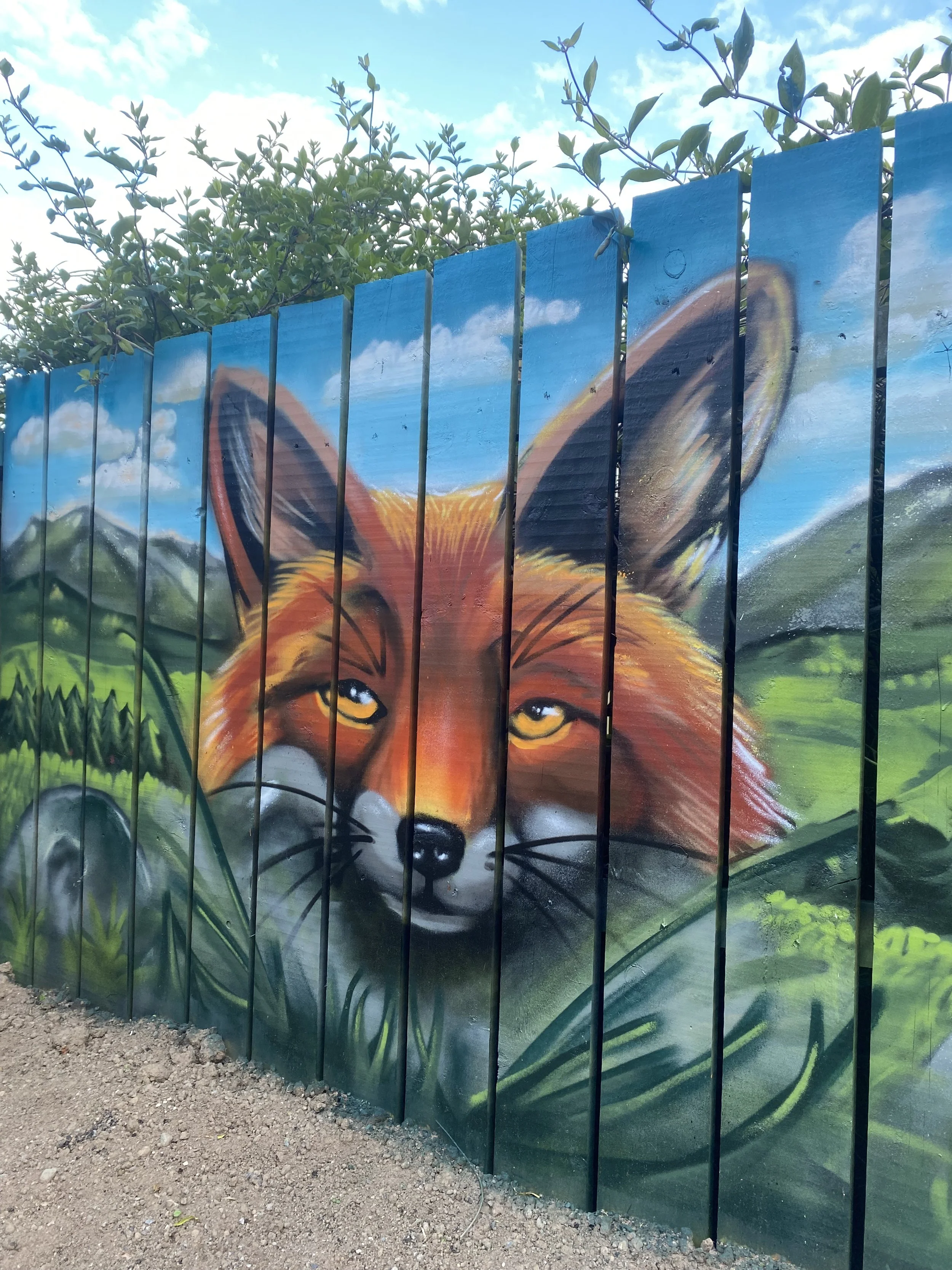 Murale peint d’un renard avec des yeux jaunes, et un paysage en arrière-plan comprenant un ciel bleu avec quelques nuages et des collines vertes.
