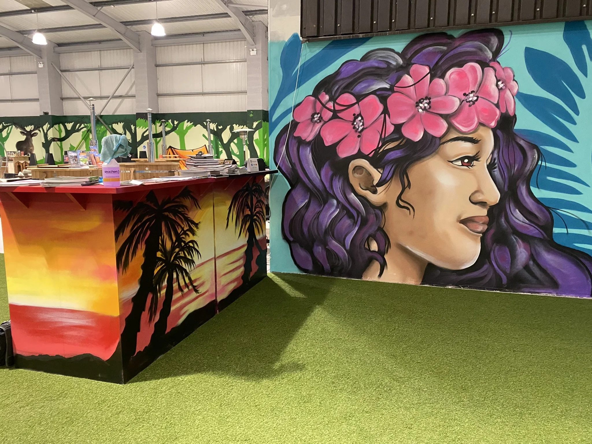 Interior of an indoor trampoline park or recreation center with colorful wall art. There is a large mural of a woman with purple hair and pink flowers in her hair, and another mural depicting a sunset with palm trees. A table with papers, a bottle of