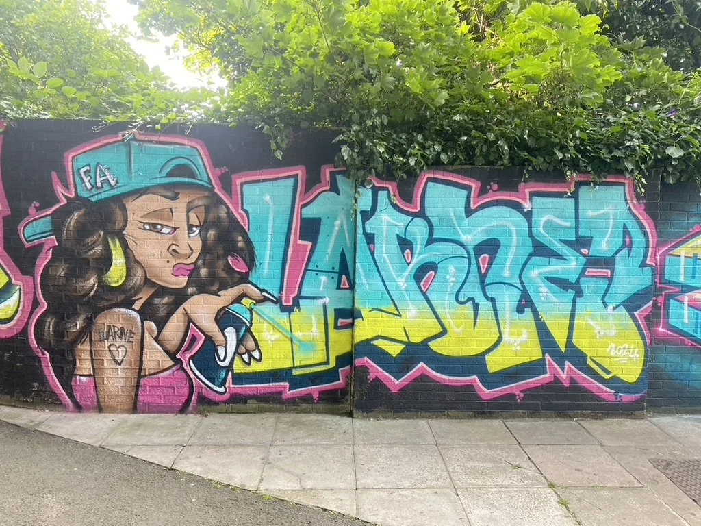 Graffiti mural featuring a woman with long, wavy hair, wearing a blue cap and yellow earrings, holding a spray paint can, with colorful stylized lettering spelling 'TURE' on a brick wall background and green foliage overhead.