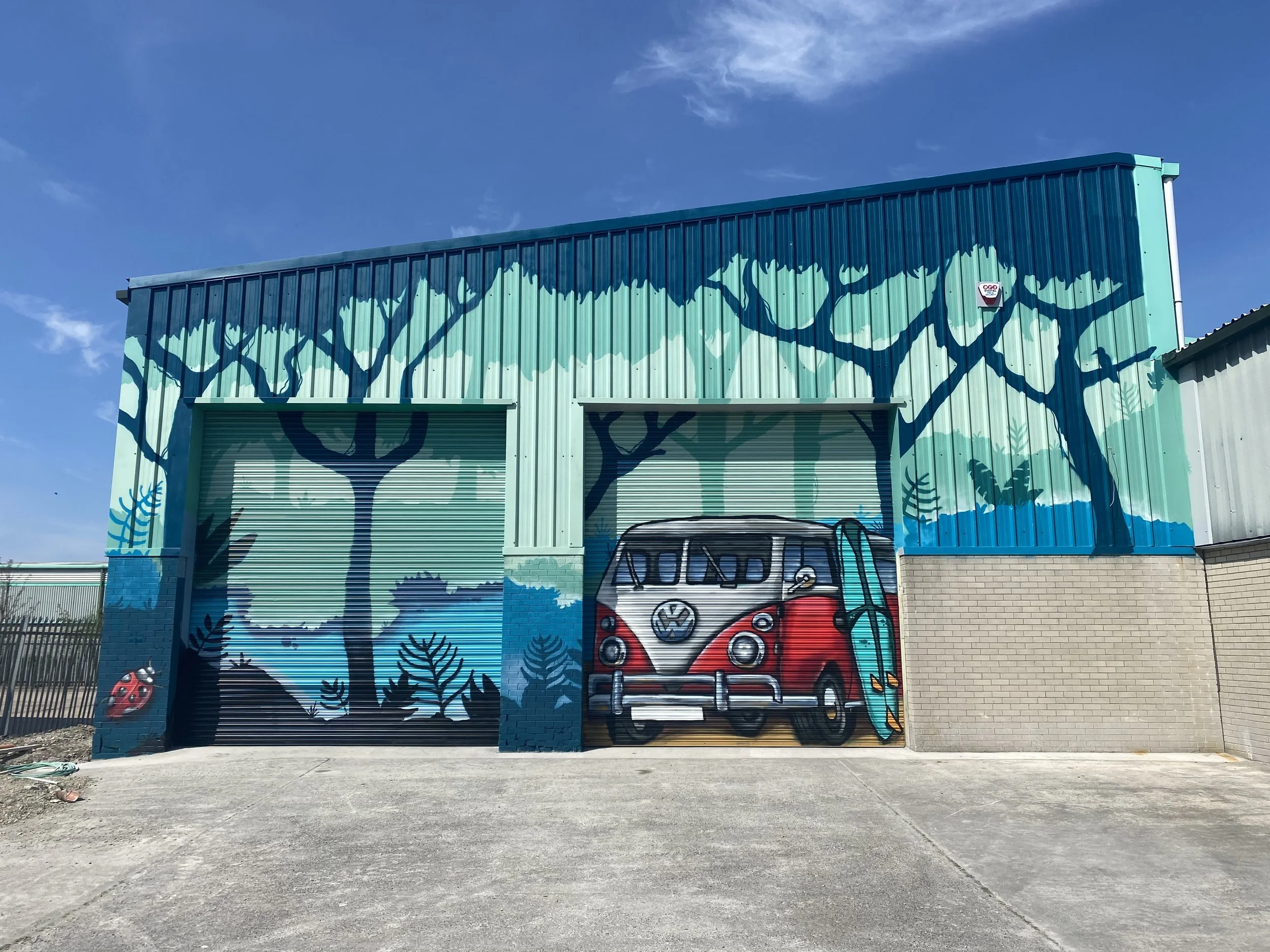 A colorful mural on a building depicts trees, a red and white Volkswagen van, ladybugs, and a ladybug and fern in the foreground, with a partly cloudy sky overhead.