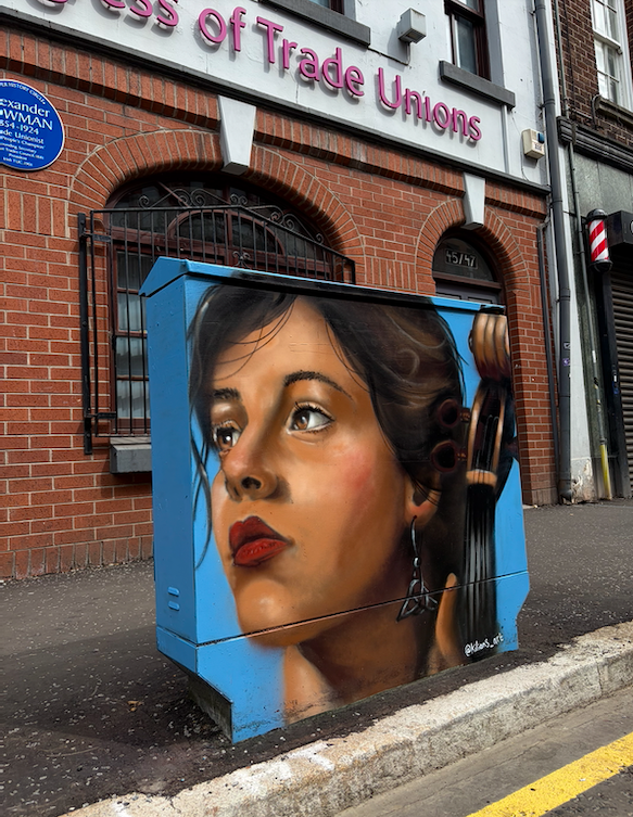 Street art graffiti of a woman with dark hair, red lipstick, and an earring, painted on a blue electrical box in front of a brick building with a sign for 'Trade Unions'.