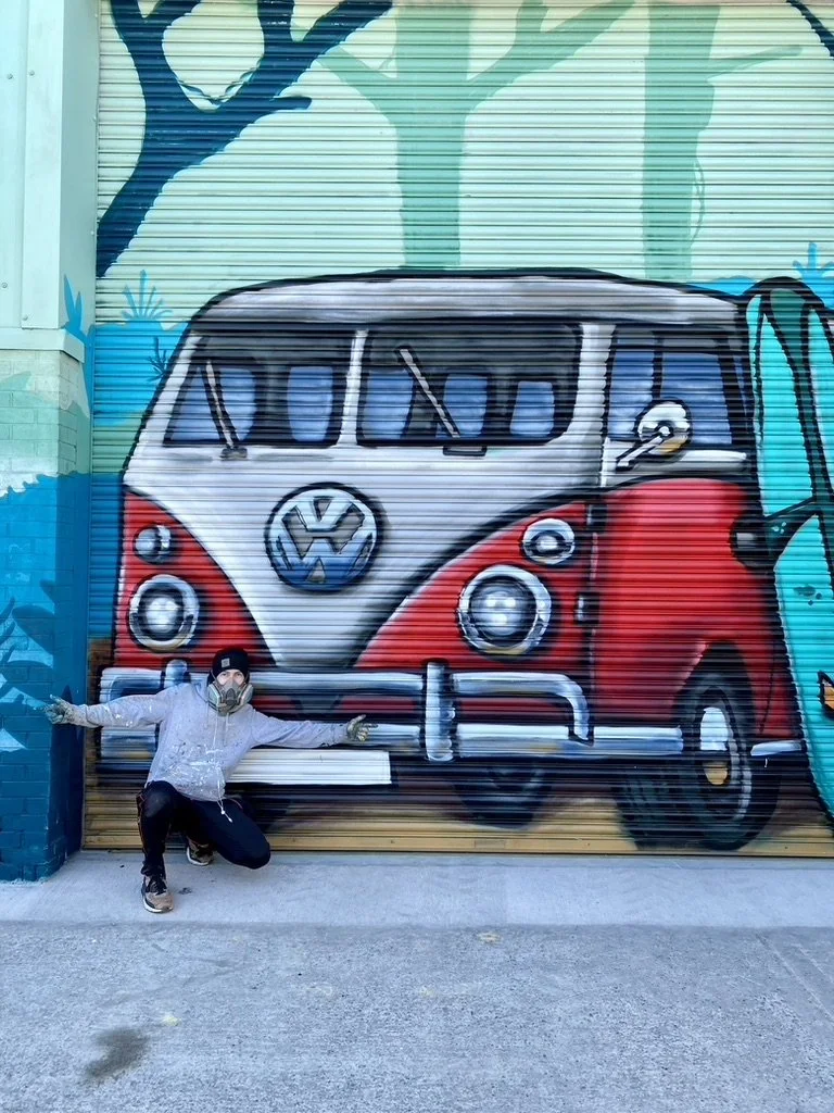 A person crouching in front of a large colorful mural of a vintage Volkswagen van. The mural is painted on a garage door and features trees in the background.