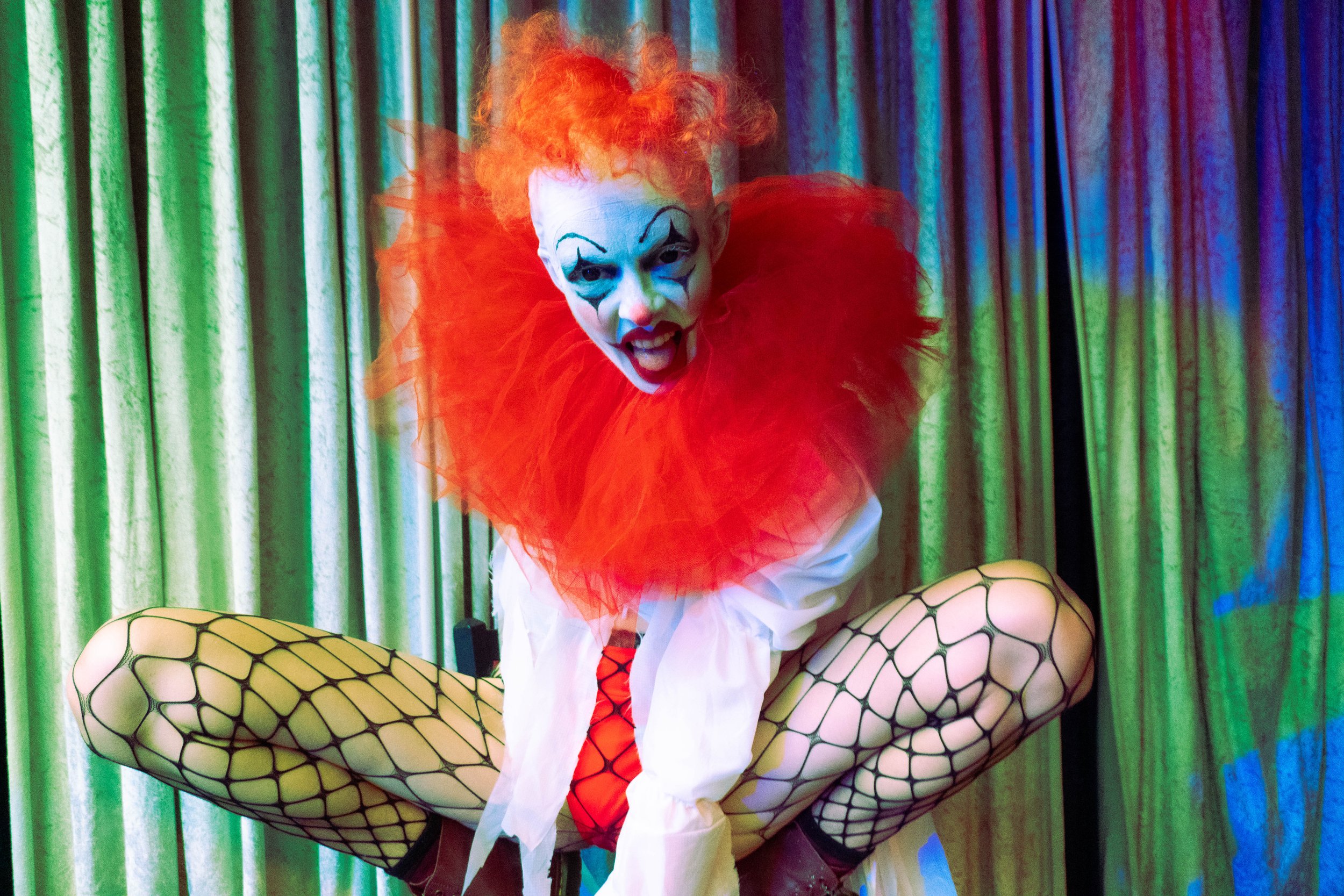 A person dressed as a clown with orange curly hair, white face paint, and exaggerated makeup, wearing a red ruffled collar and fishnet stockings, sitting in front of a colorful curtain.