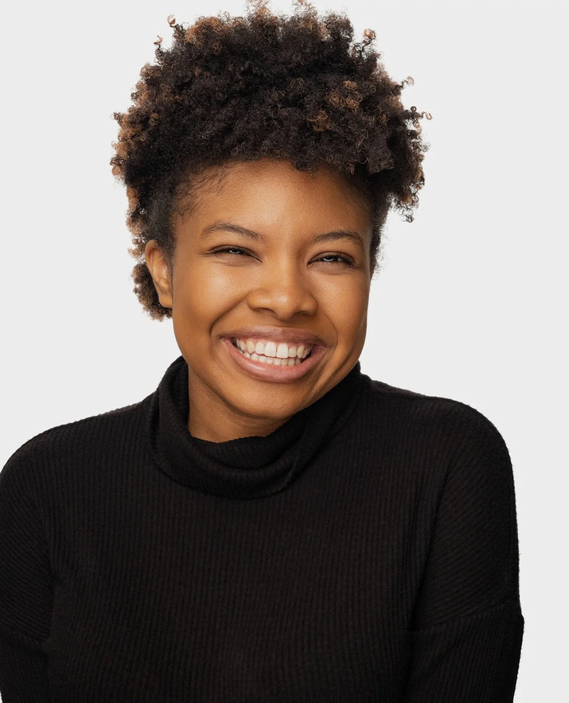 A smiling woman with short curly hair wearing a black turtleneck sweater, posing against a plain white background.