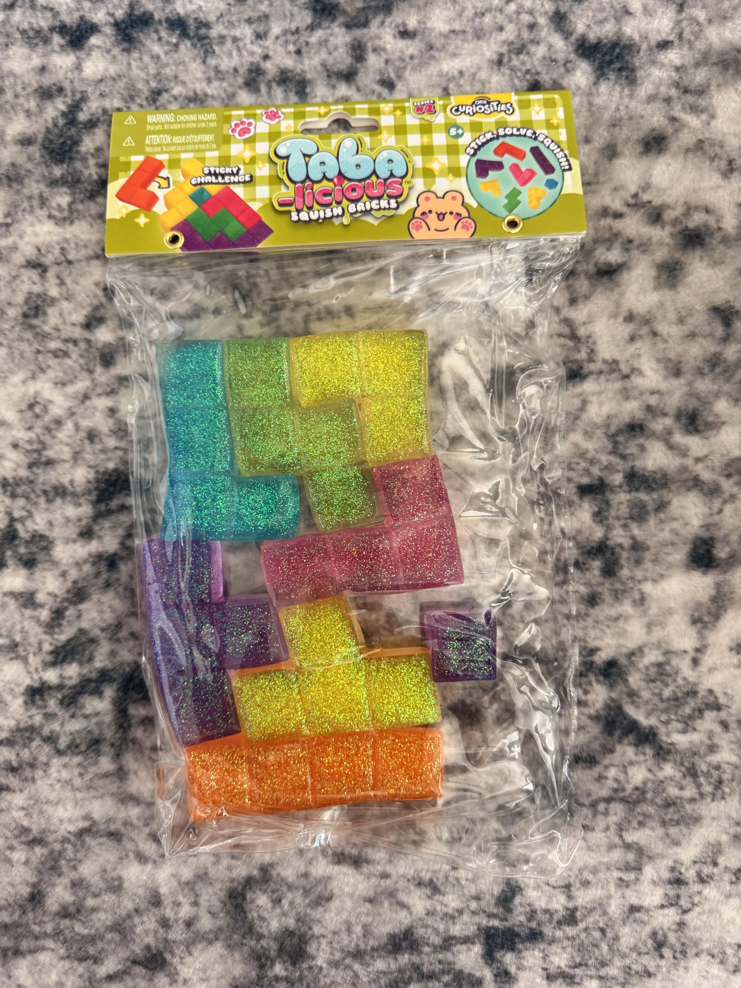 Packaged set of colorful, glittery slime bricks in various colors, in a plastic bag with a bright green and yellow checkered label featuring cartoon animal characters and puzzle images.