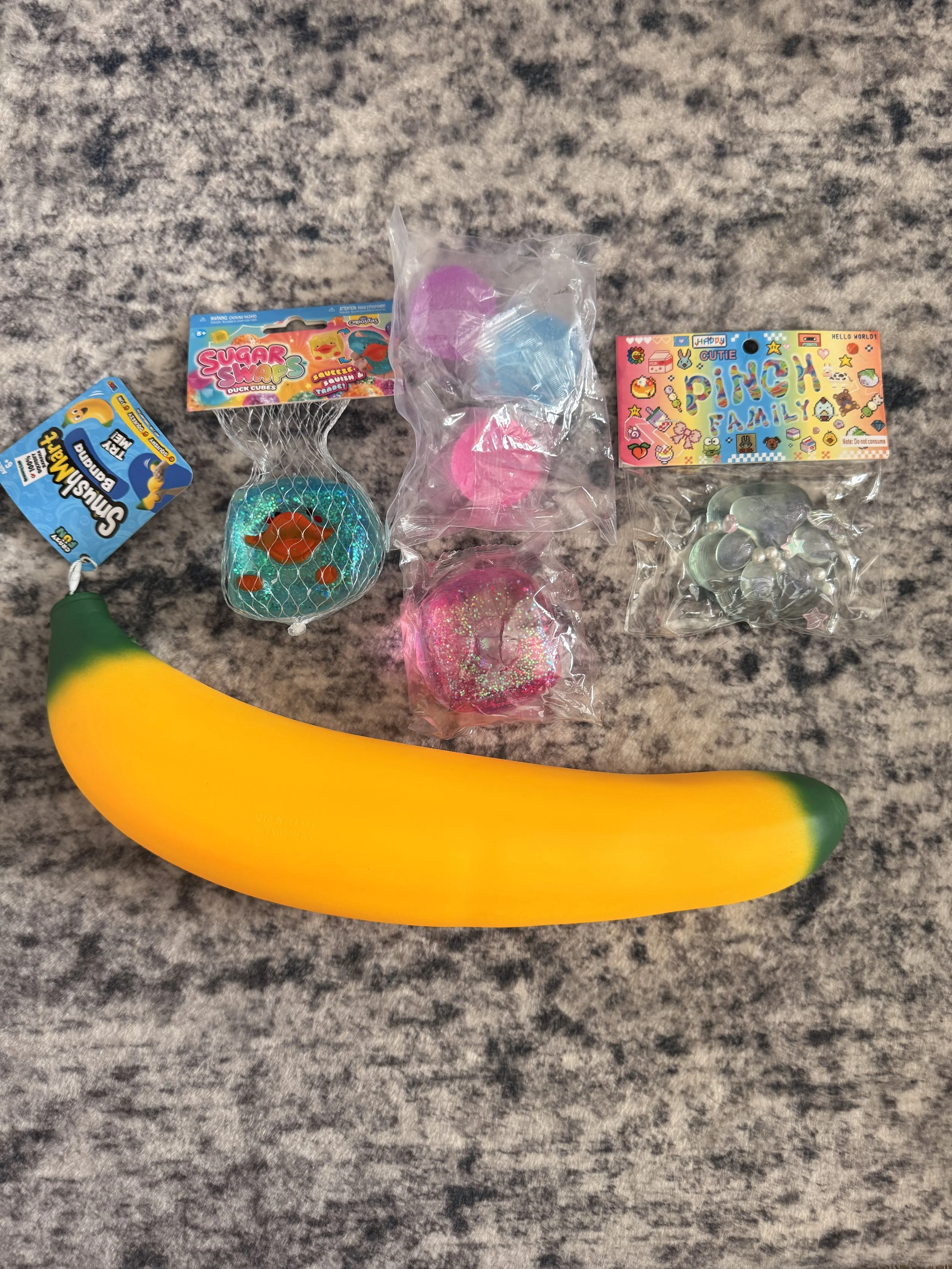 Colorful foam bath bombs, a yellow and green rubber banana, and toy packaging on a gray textured surface.