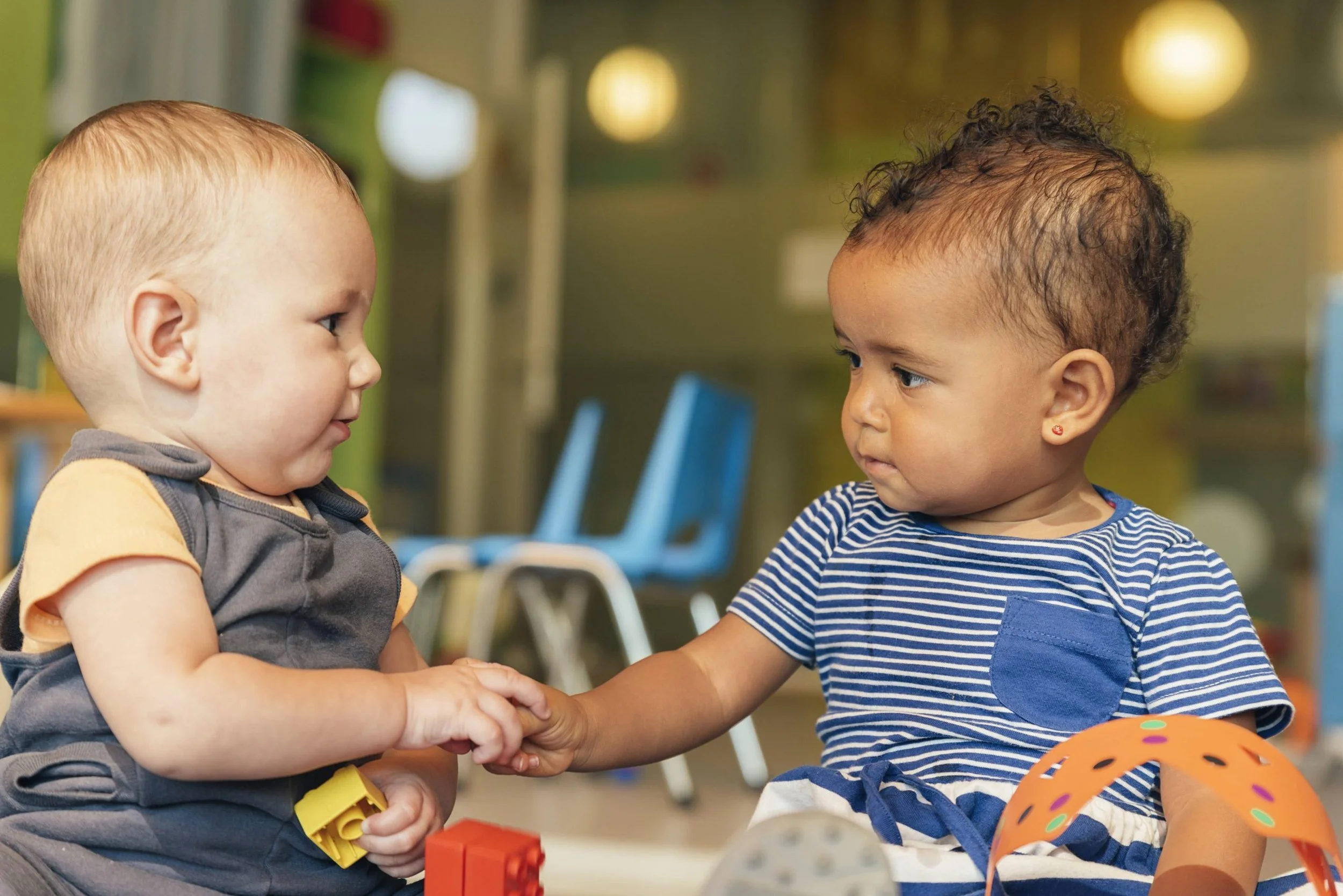 Two young children, a blonde boy and a curly-haired girl, are holding hands and looking at each other in a colorful room, possibly a classroom or daycare, with toys and chairs in the background.