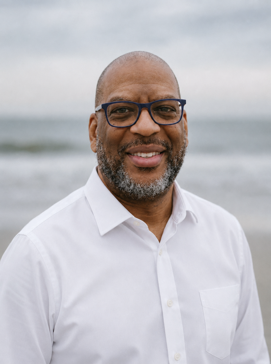 A smiling Black man with glasses and a beard wearing a white shirt standing on a beach with the ocean in the background.