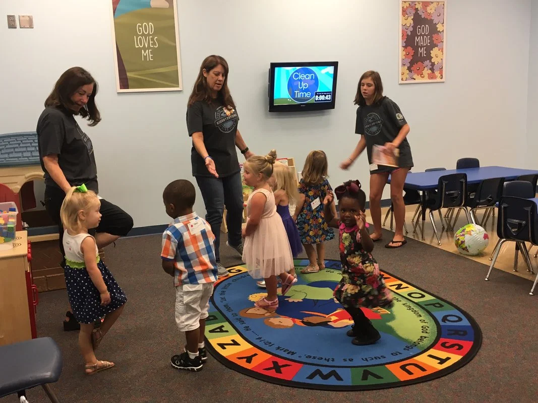 Young children participating in a preschool activity with teachers in a classroom, standing on an alphabet-themed rug, with a television screen displaying 'Clean Up Time' in the background.