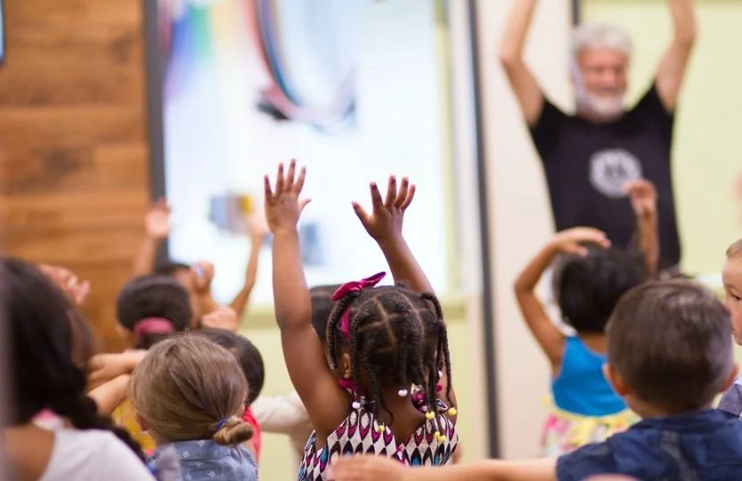 Children in a classroom with raised hands, listening to a teacher or speaker at the front.