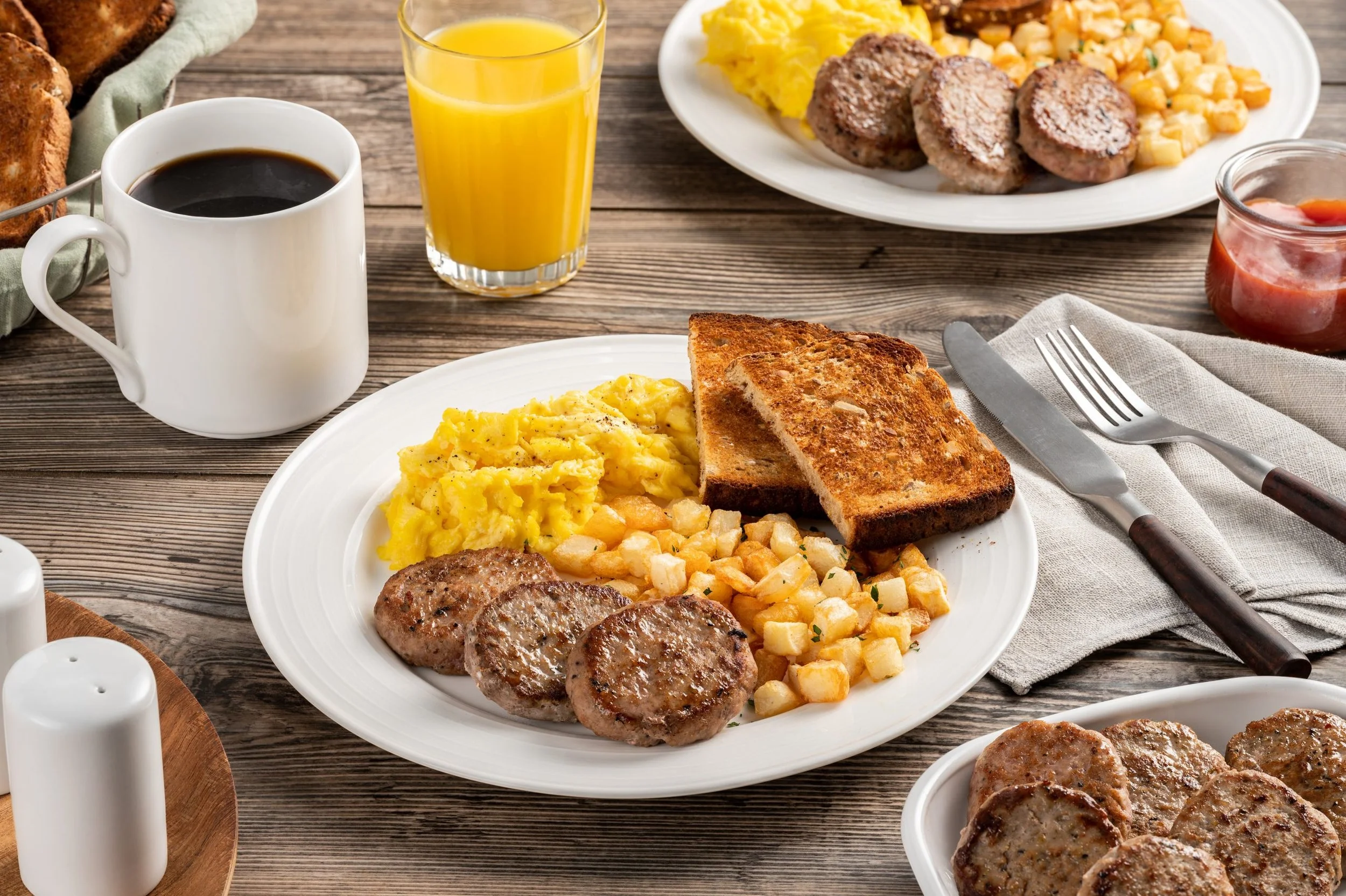A breakfast meal on a wooden table including scrambled eggs, sausage patties, toasted bread, hash browns, a glass of orange juice, a cup of coffee, a bowl of ketchup, and a plate with scrambled eggs, sausage patties, hash browns, and toast.