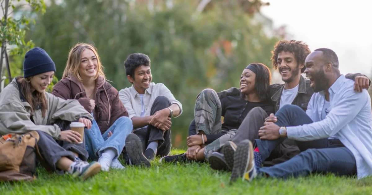 Group of six friends sitting on grass, smiling, and enjoying outdoor weather in a park.