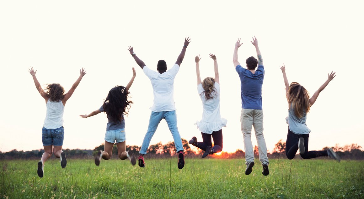 Group of six young people jumping in the air on a grassy field at sunset.