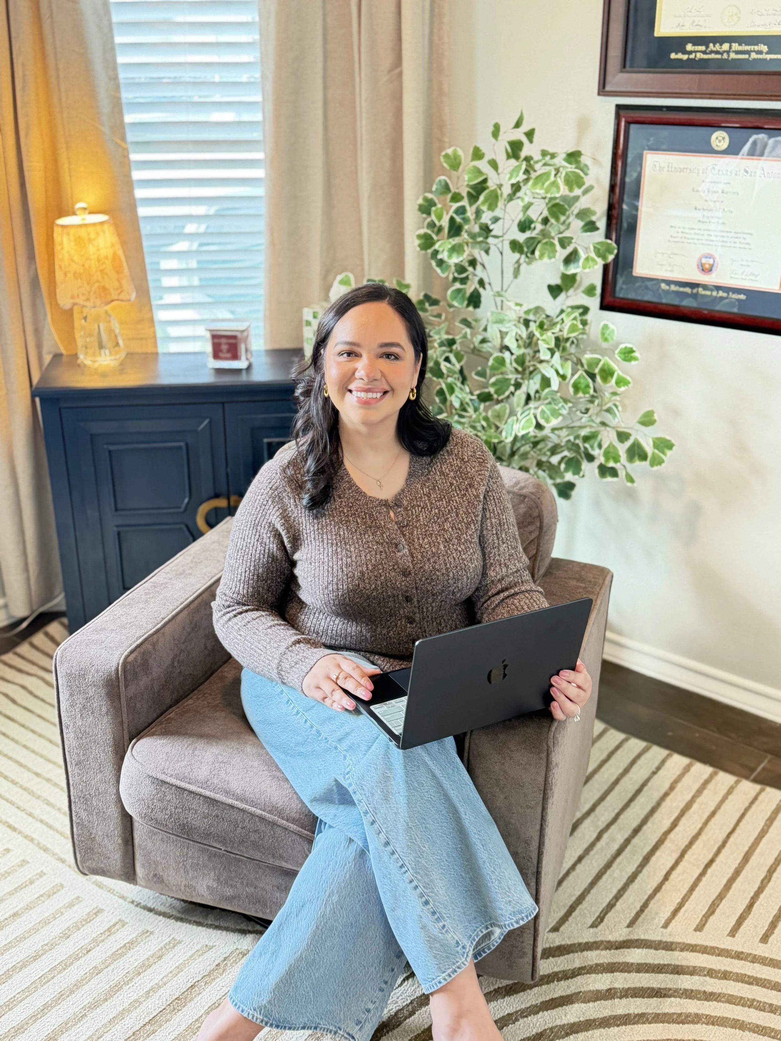 A woman sitting on a beige armchair, working on a laptop, in a room with striped carpet, a black cabinet, and a large plant behind her. There are framed certificates on the wall and a window with blinds.