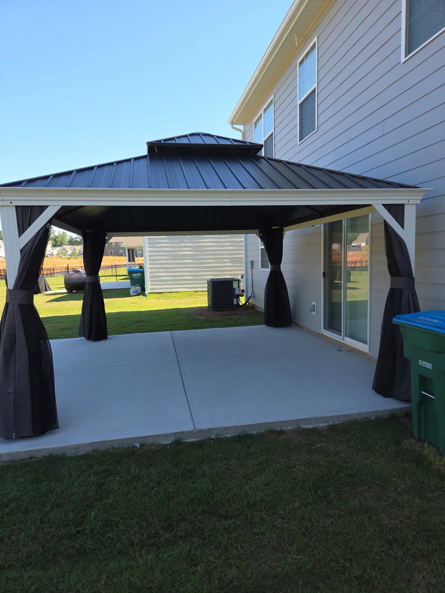A backyard with a covered patio, black curtains, green grass, a trash bin, and an air conditioning unit, with a house in the background.