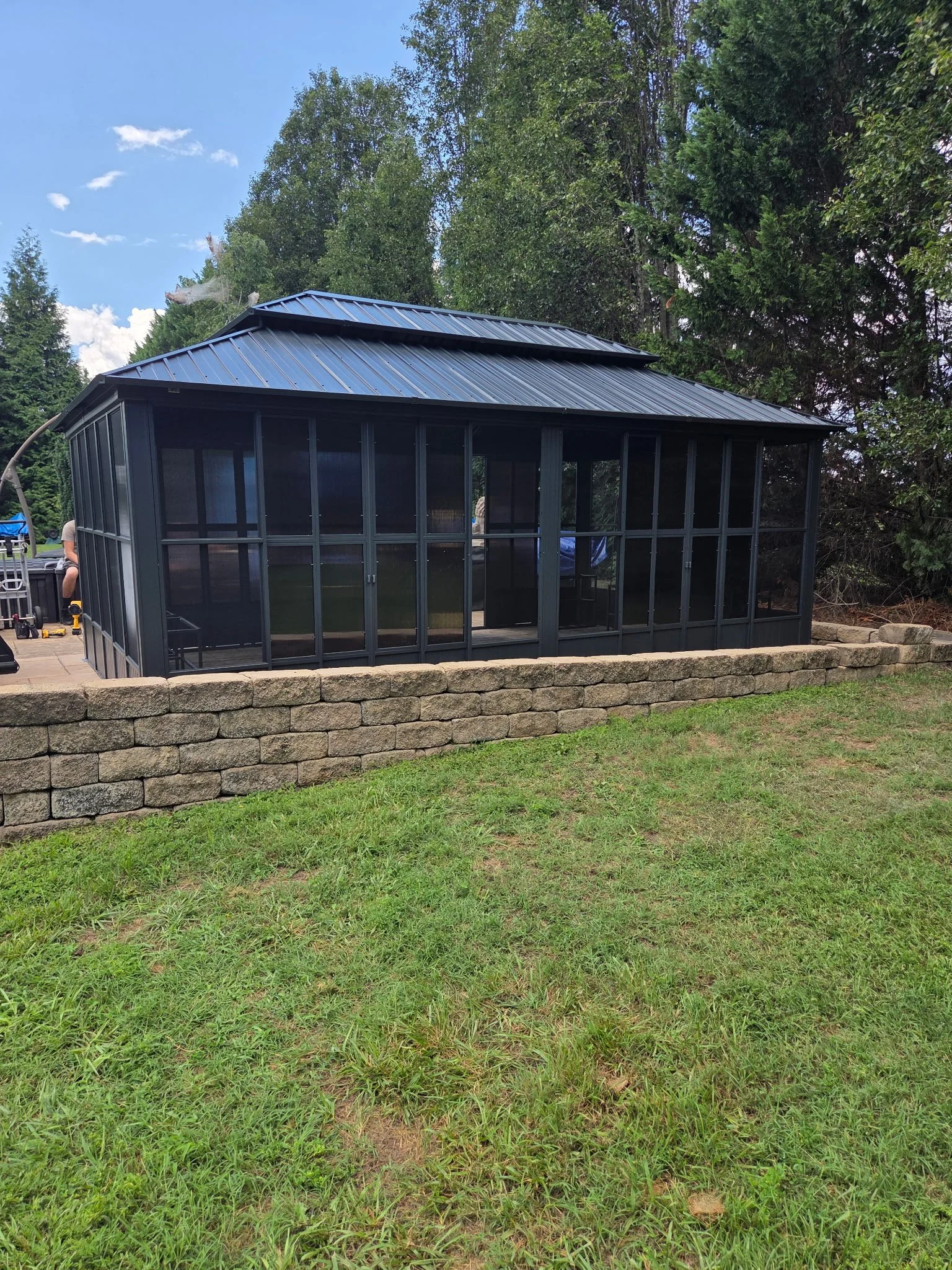 A screened outdoor structure with a metal roof, situated on a grassy lawn with trees in the background.