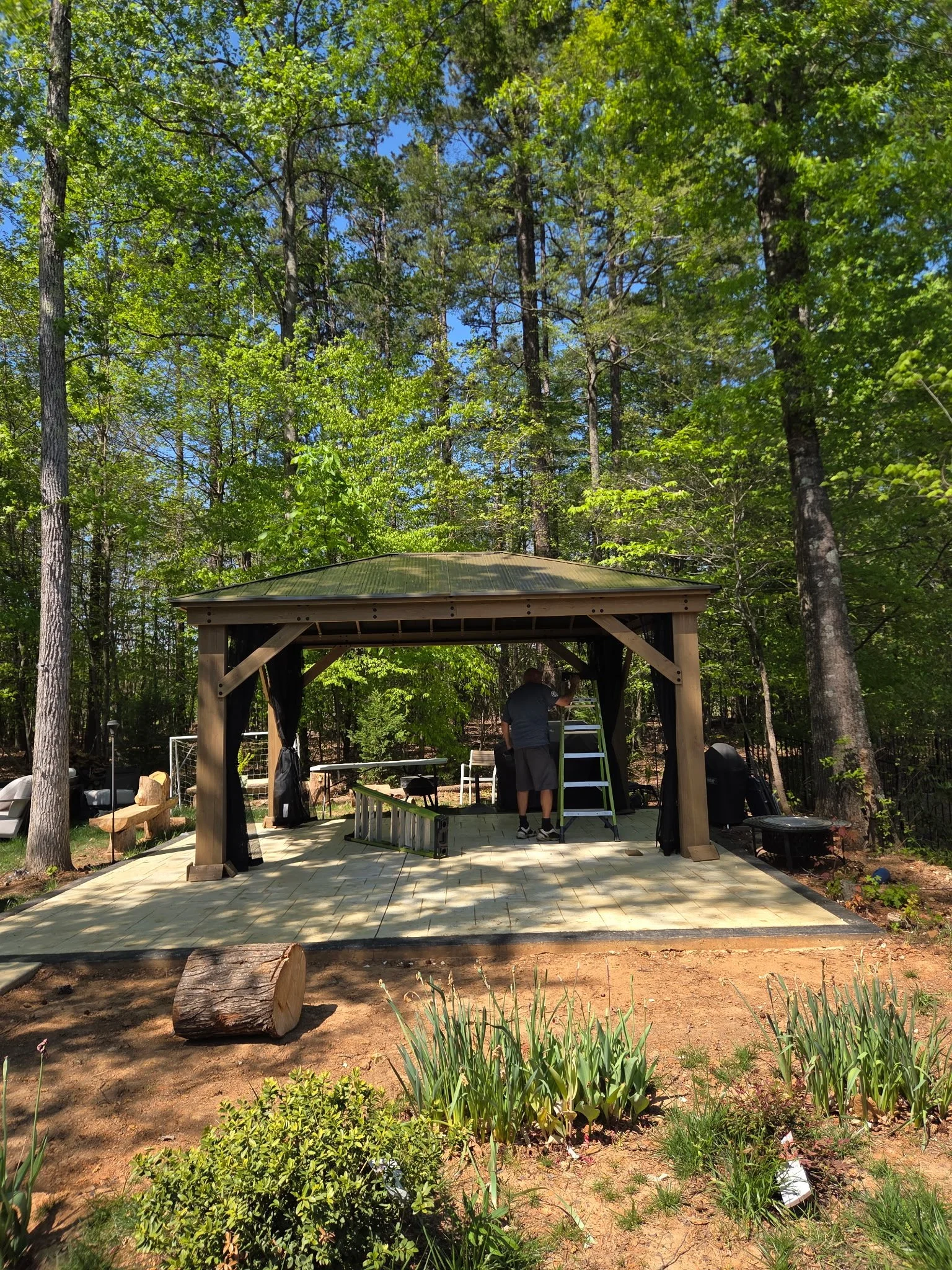 A person working on constructing or repairing a wooden gazebo in a lush, green backyard. The gazebo is on a paved patio with gardening tools and supplies nearby, surrounded by trees and plants.