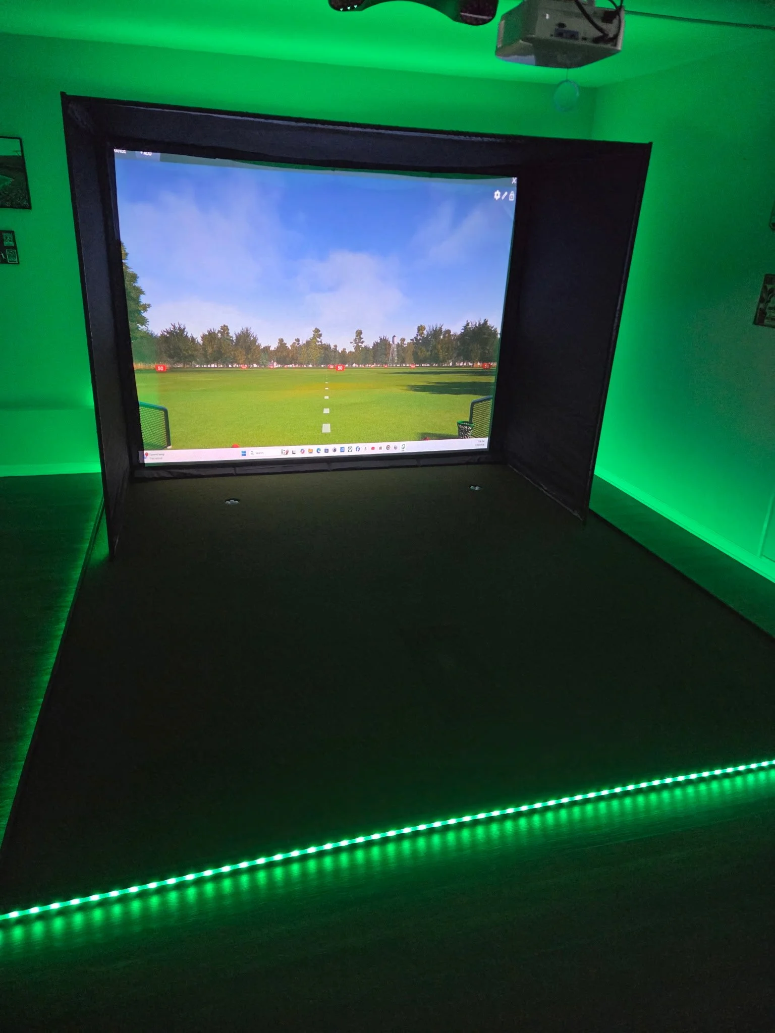 An indoor golf simulator setup with a large screen displaying a golf course and green lighting along the edges of the platform.