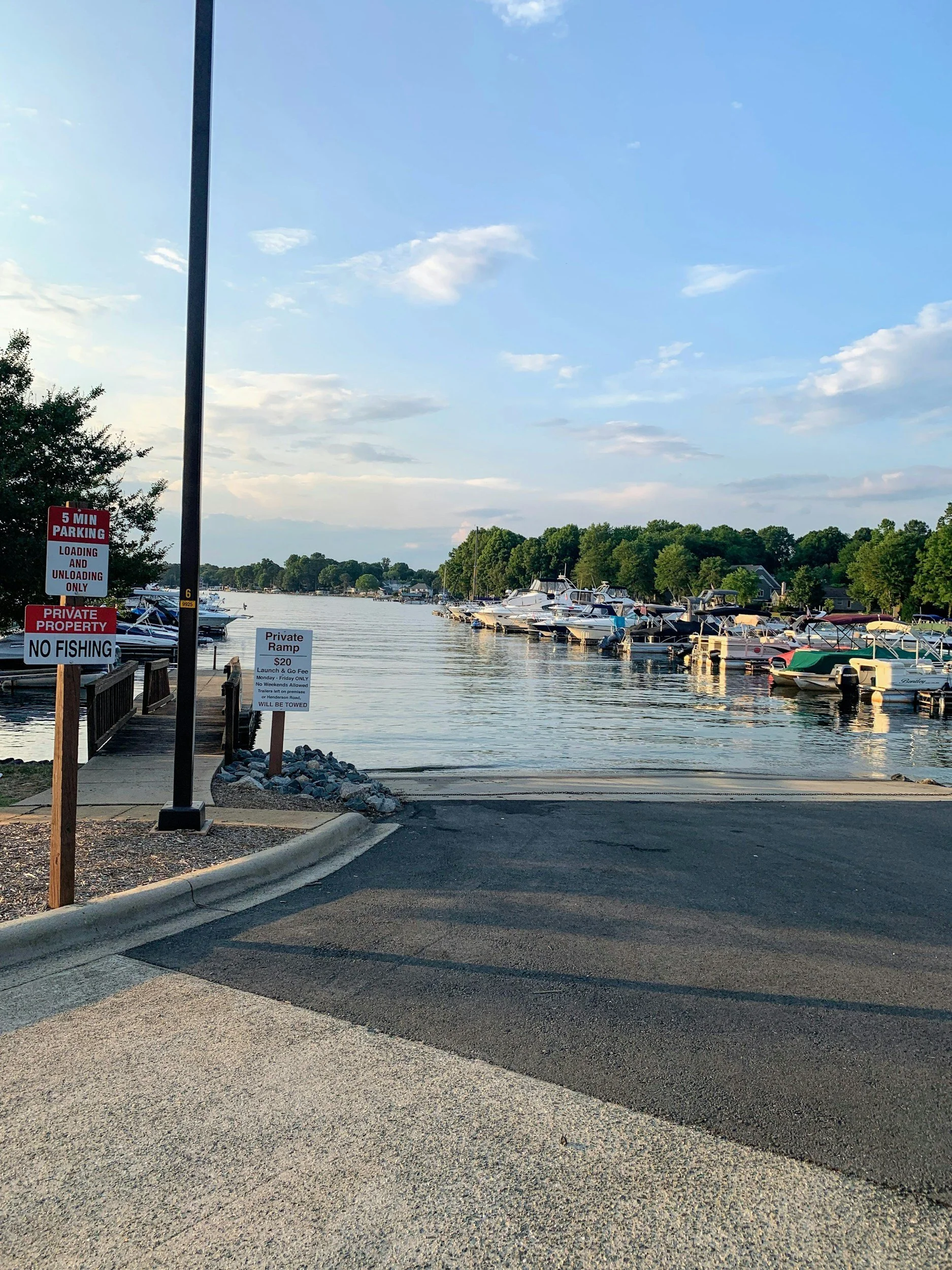 View of a marina with boats docked along the water, a sign indicating no fishing, and a clear sky with some clouds.