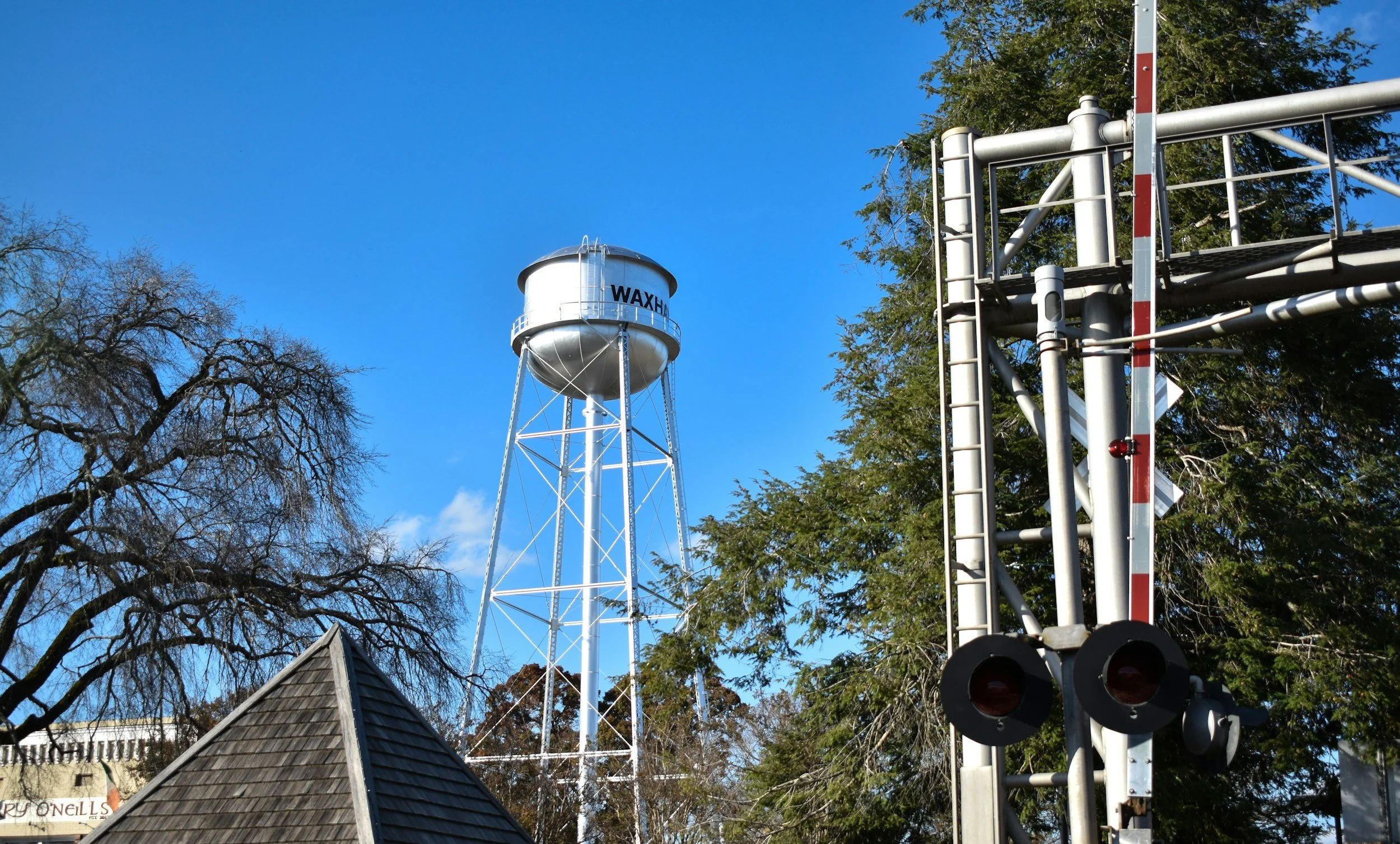 A white water tower labeled 'WAXHAW' in black letters. To the right, there is a metal structure with pipes and red and white striped barriers. There are trees with some bare branches and some green leaves, and a building with a sloped roof in the foreground.