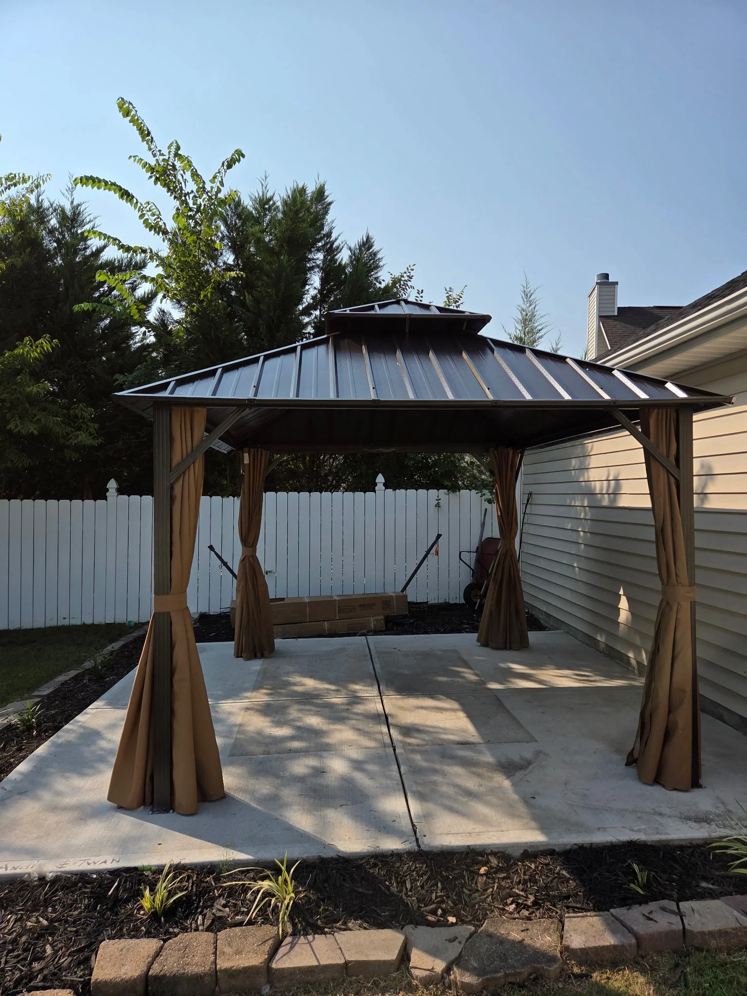 Backyard patio with a gazebo, white fence, garden bed, and house in the background, with trees and clear sky.