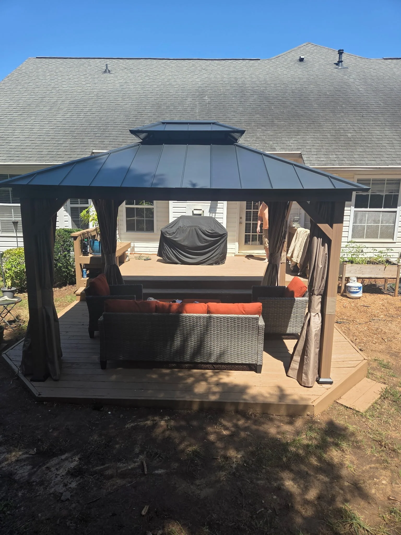 A backyard deck with a gazebo and seating area under a covered pergola. The background shows a house with gray shingles and a clear blue sky.
