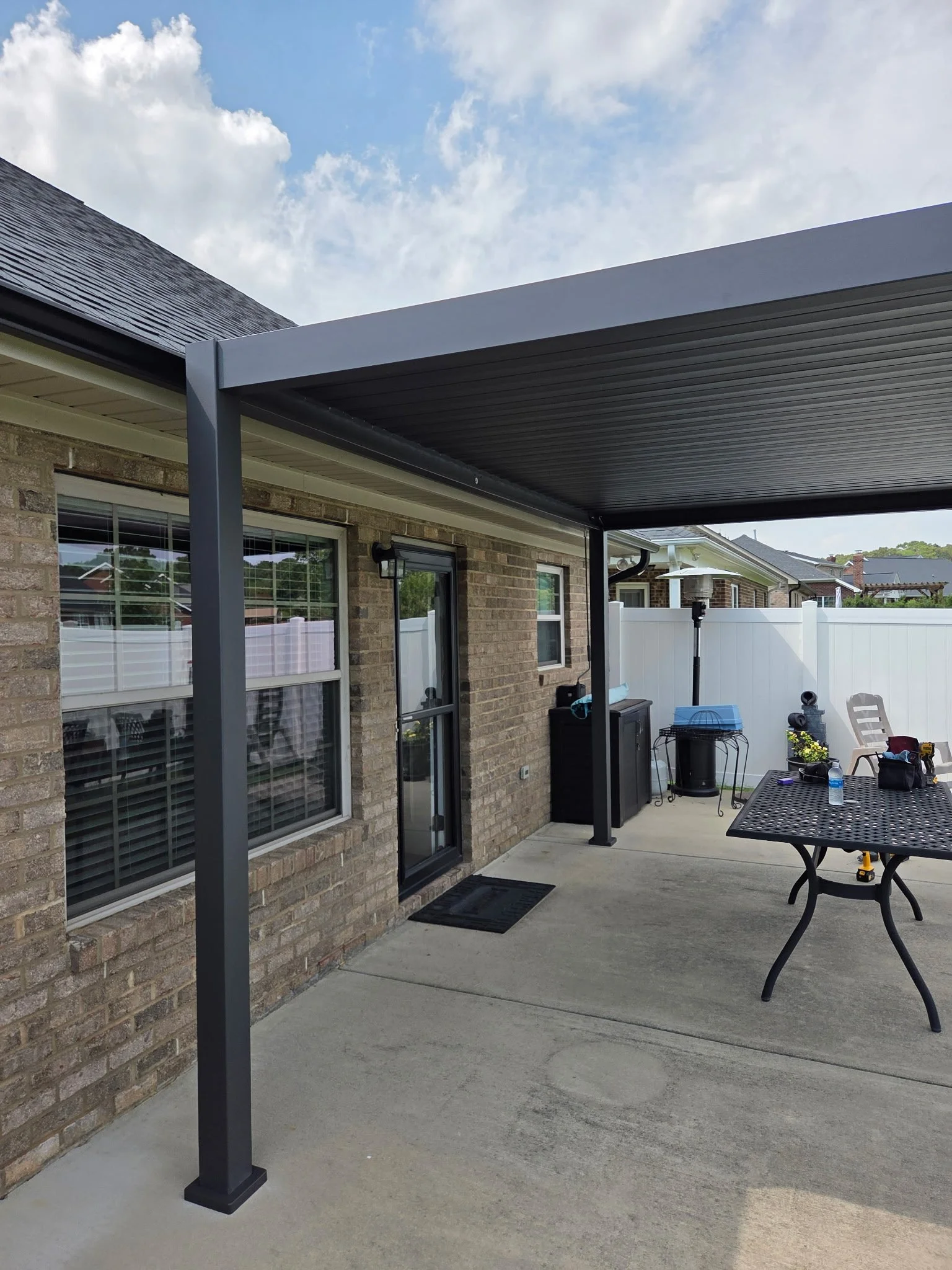 Backyard patio with a brick house wall, two large windows with white blinds, a black door, a black roof-covered patio area, an outdoor table, a water bottle, and various decorations. White fence in the background, cloudy sky.