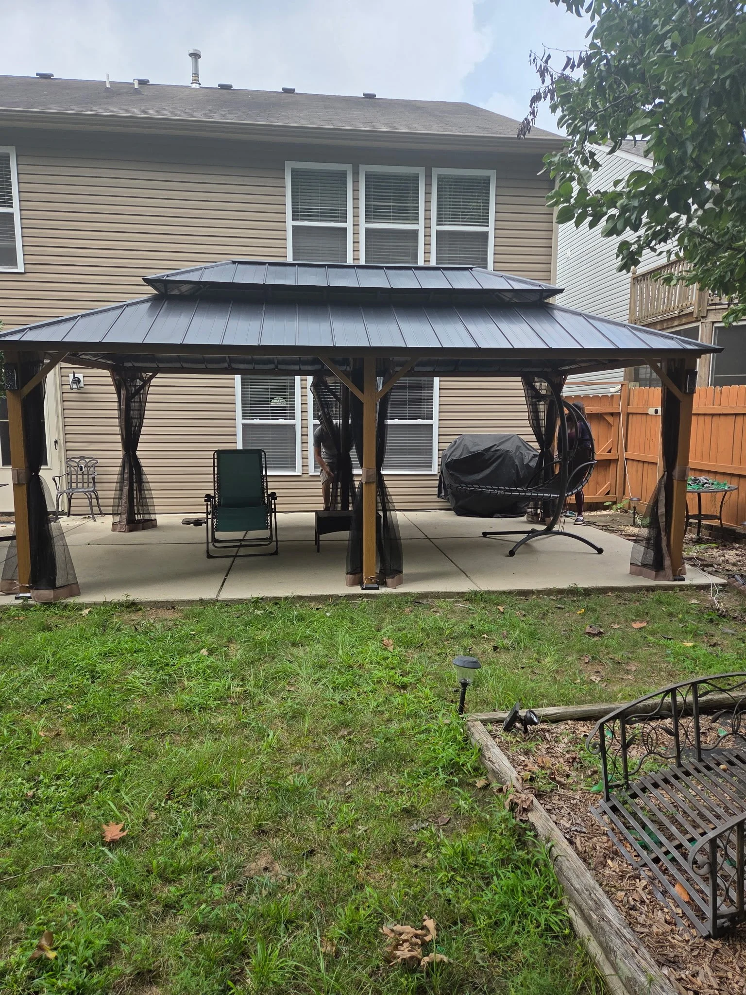 Backyard patio with a metal gazebo, outdoor chairs, a grill covered with a tarp, and a grassy lawn bordered by a wooden fence.