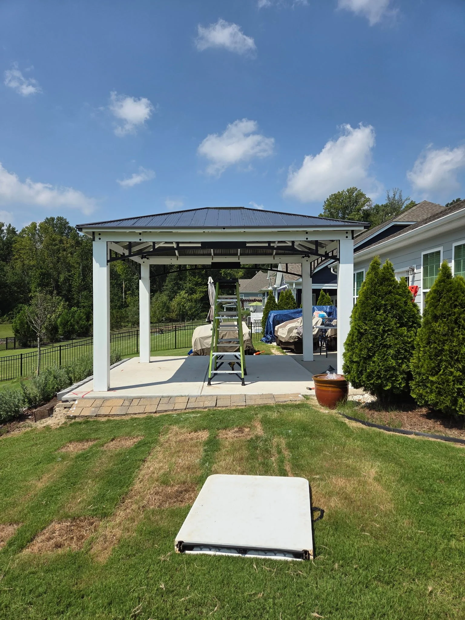 A backyard with a covered patio structure, a ladder, covered items, and a small white table on a grassy lawn under a partly cloudy sky.