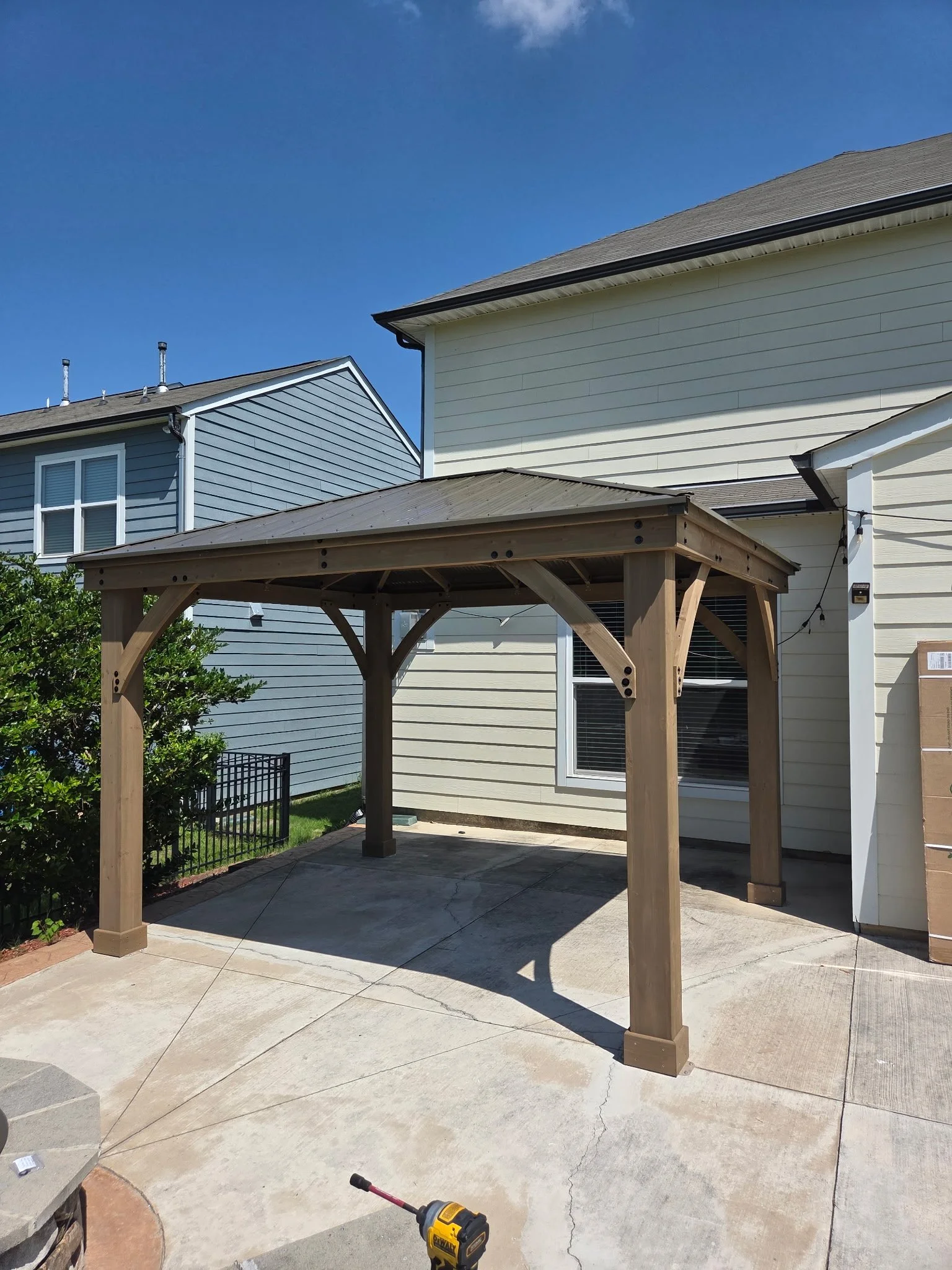 The image shows a backyard with a newly constructed wooden pergola over concrete patio, situated near a house with white and light blue siding. There is a small green bush on the left, a string of outdoor lights on the right side of the house, and a 