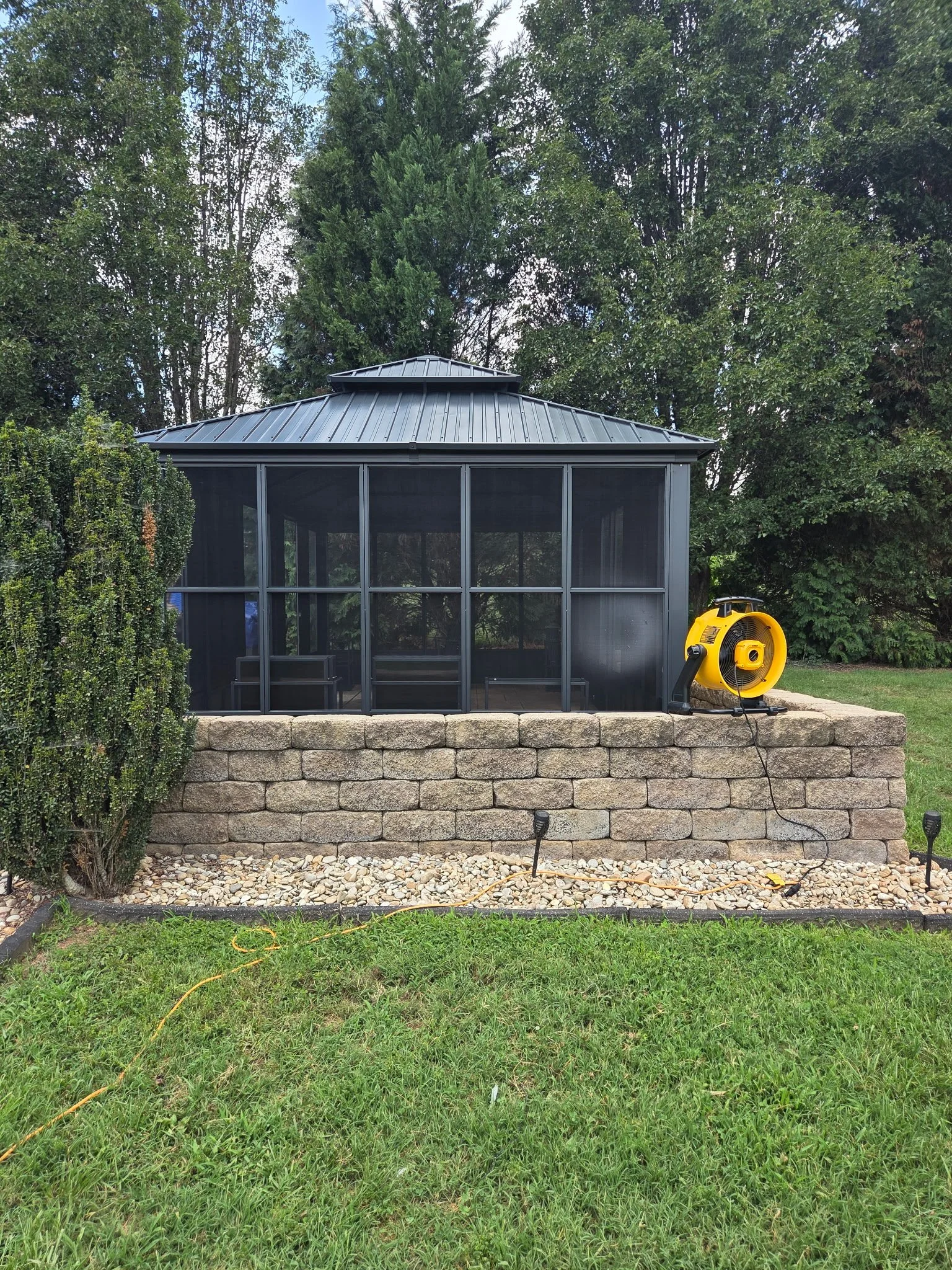 A screened gazebo with a metal roof on a stone block wall, surrounded by green grass and trees, with a yellow outdoor fan or heater placed on the right side.