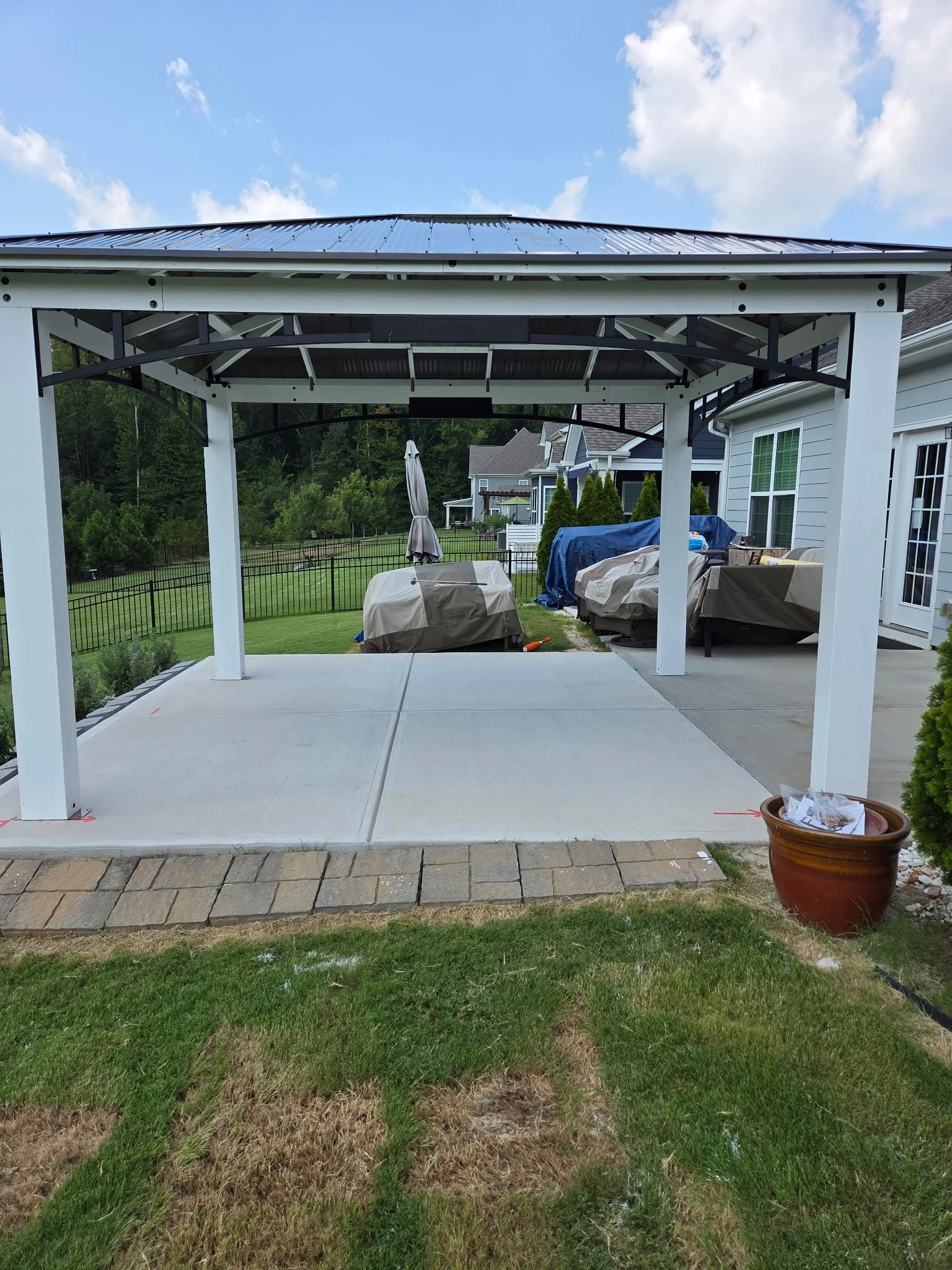 Backyard patio with new concrete slab, covered porch with black metal roof frame, covered outdoor furniture, plants, and a backyard with green lawn, trees, and neighboring houses.