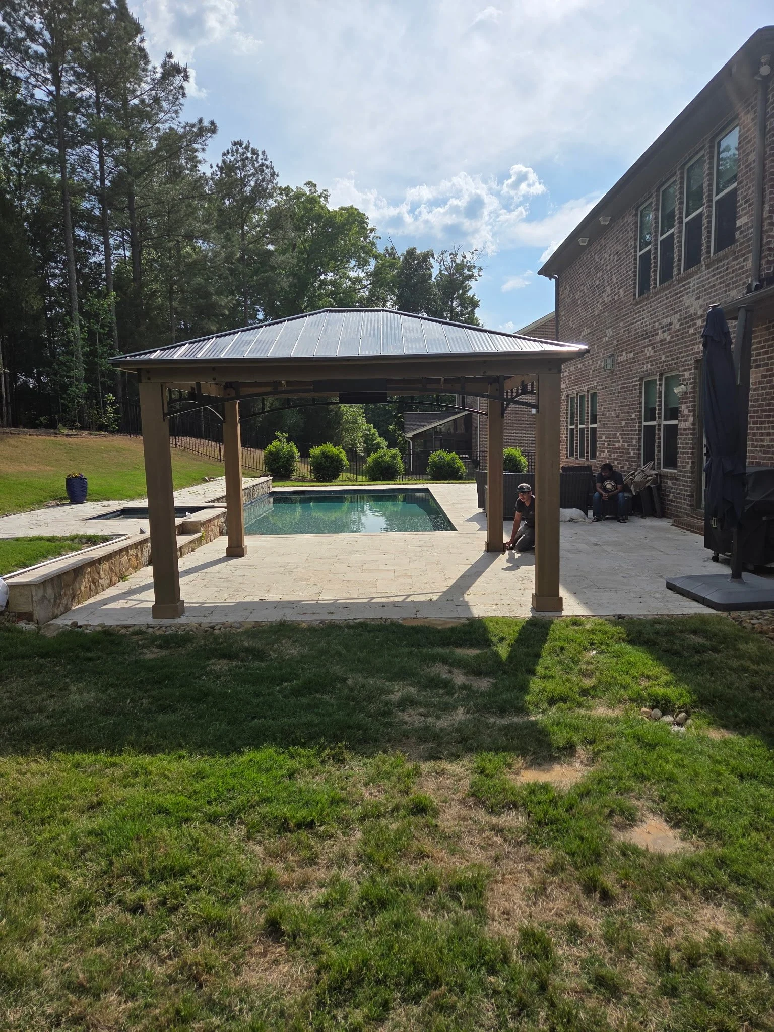 A backyard with a small pool, a stone patio, a covered pergola, and a brick house with multiple windows. There are trees in the background and a person near the pool.