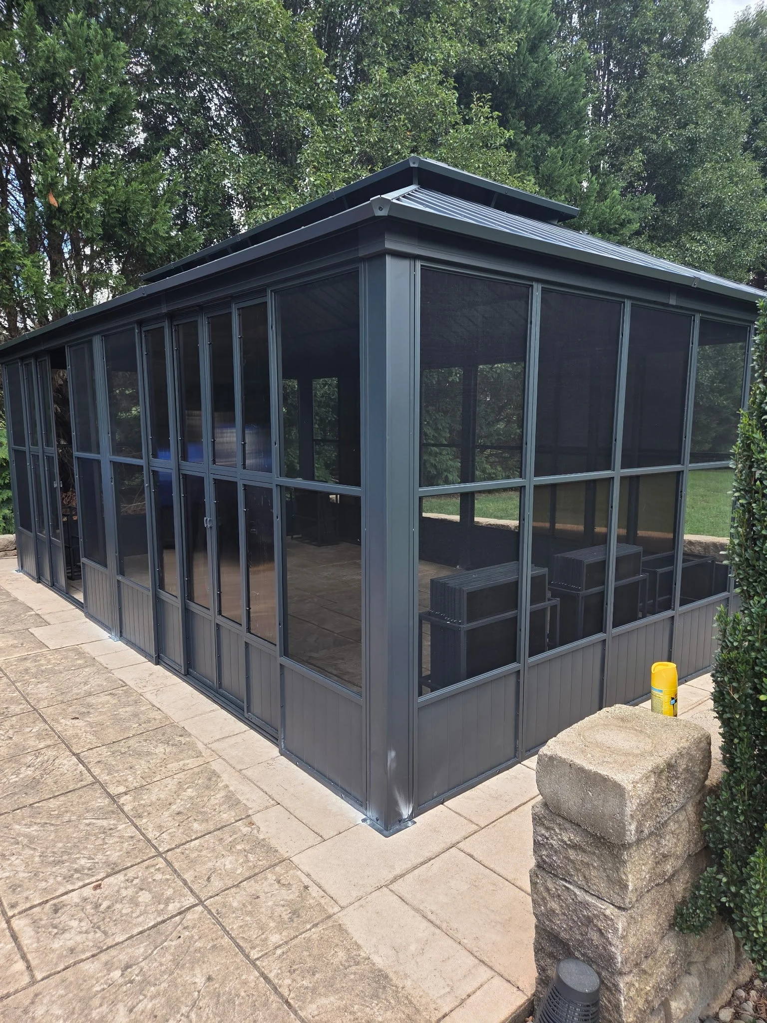 A screened outdoor patio or sunroom with large glass windows and a metal roof, situated on a stone patio with a garden in the background.
