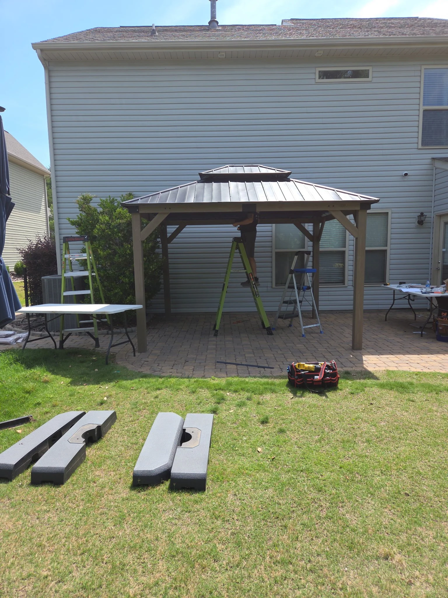 Person installing a metal roof on a gazebo in a backyard. Tools and ladders are present, with a house in the background.