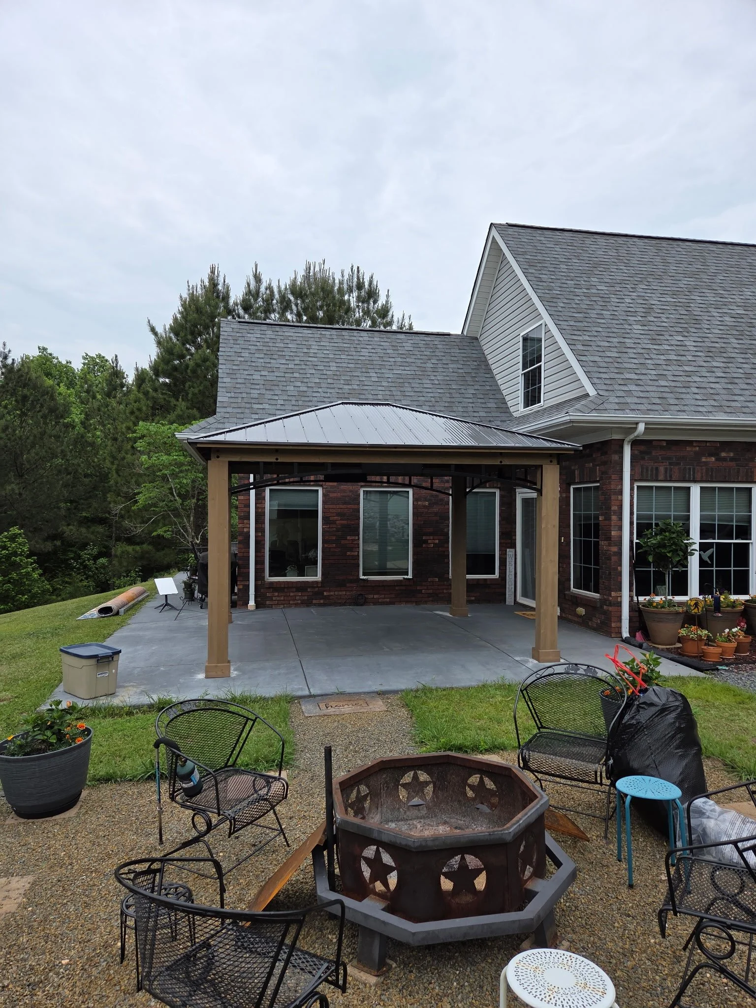 View of a backyard patio area with garden chairs, a fire pit, potted plants, a wooden pergola, and a house with a brick exterior and large windows, under a cloudy sky.