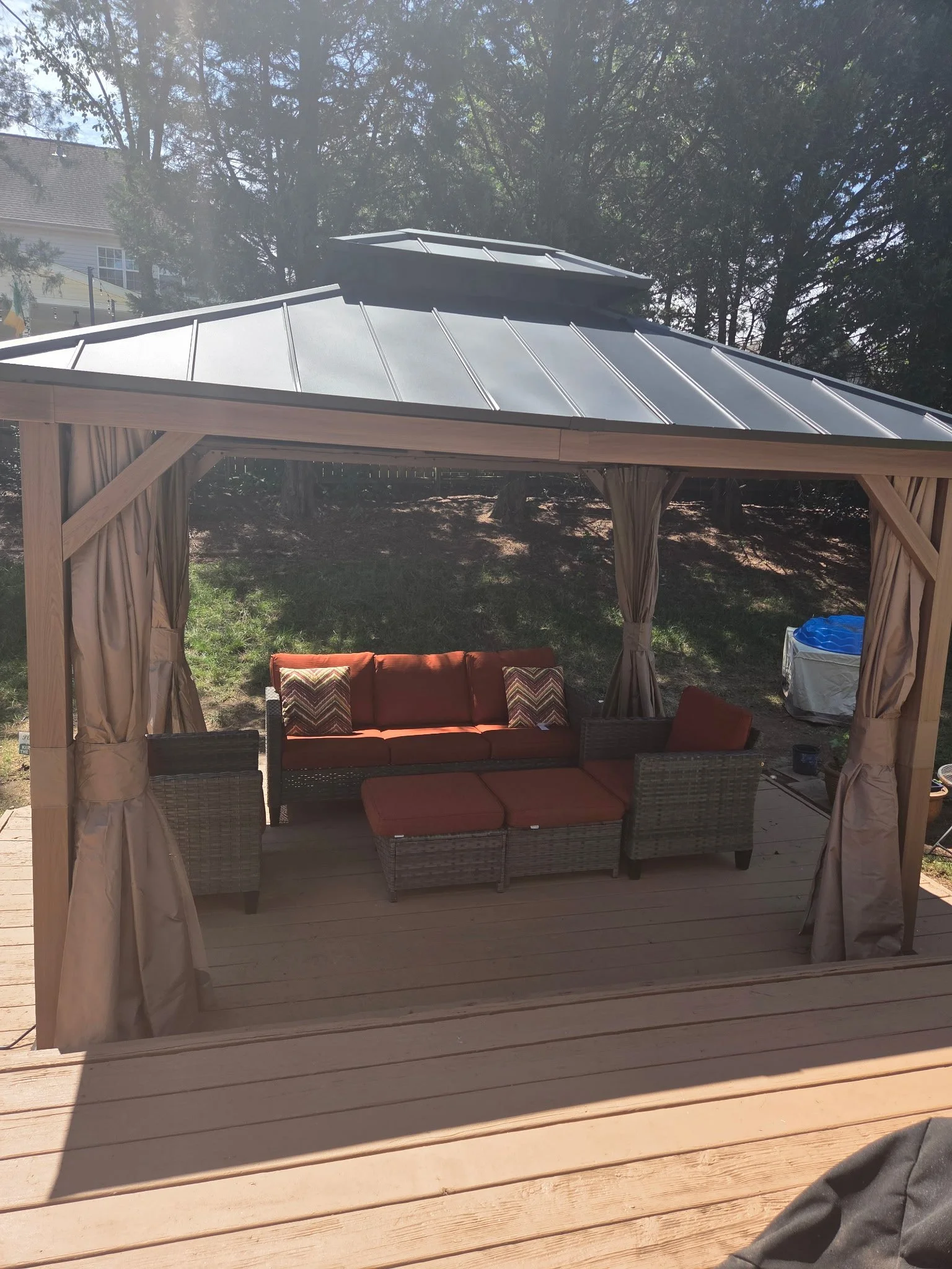 View of a wooden outdoor gazebo frame with curtains, an orange cushioned outdoor sofa with patterned pillows, and a wooden deck, with trees and a house in the background.