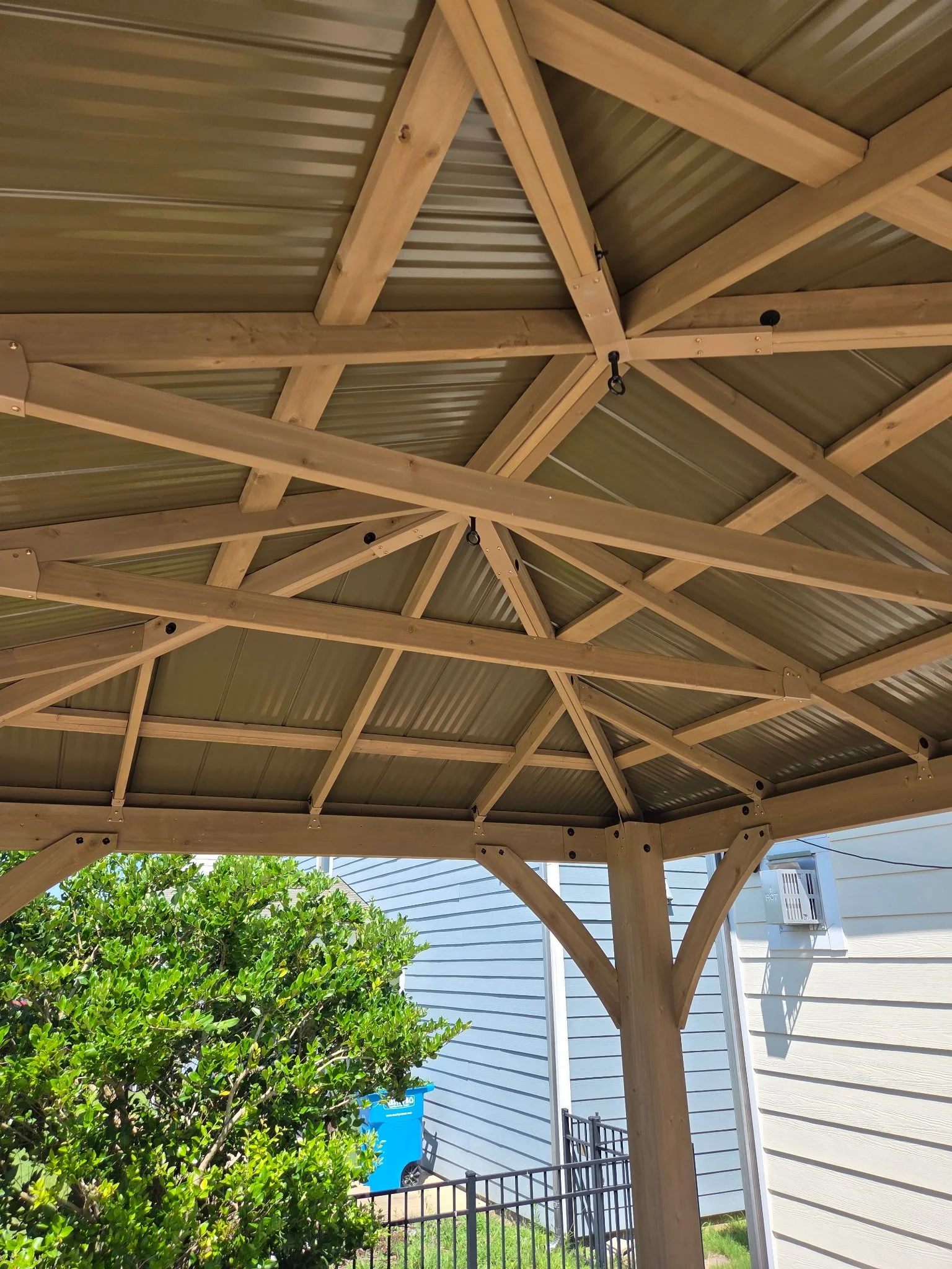 The image shows the underside of a wooden gazebo roof with a metal roof sheet. There are support beams and cross braces with metal hardware, and a metal hook hanging from one of the beams. In the background, there is green foliage, a blue recycling b