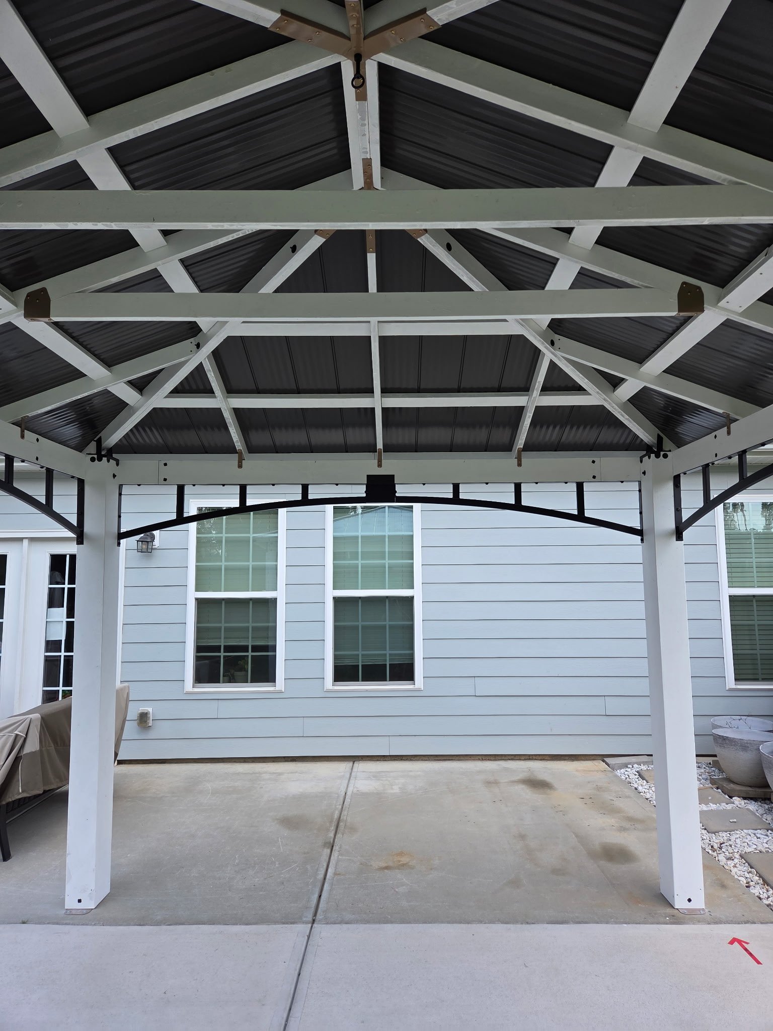 The image shows a covered outdoor patio area with a white wooden pergola or frame that has a dark metal roof. In the background, there is a house with light blue siding, large windows, and white trim. The patio has a concrete floor, and on the right 