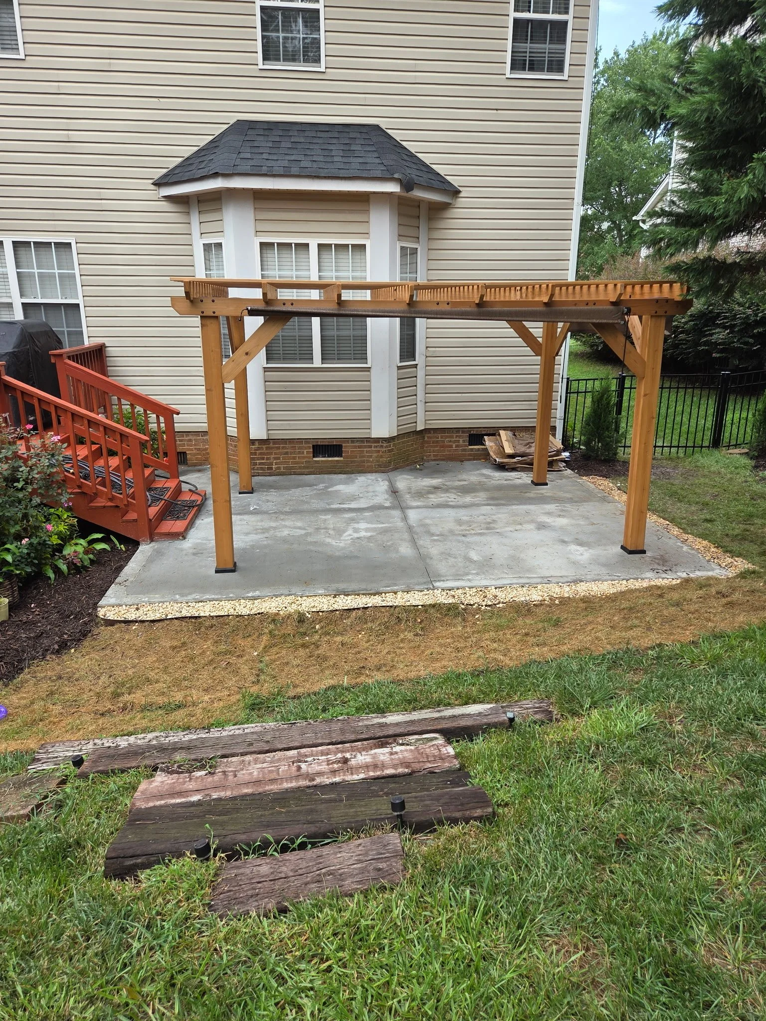 Backyard with a partially built wooden deck structure, a concrete patio, and a garden with bushes and flowers. There is a house in the background with beige siding and white window frames.