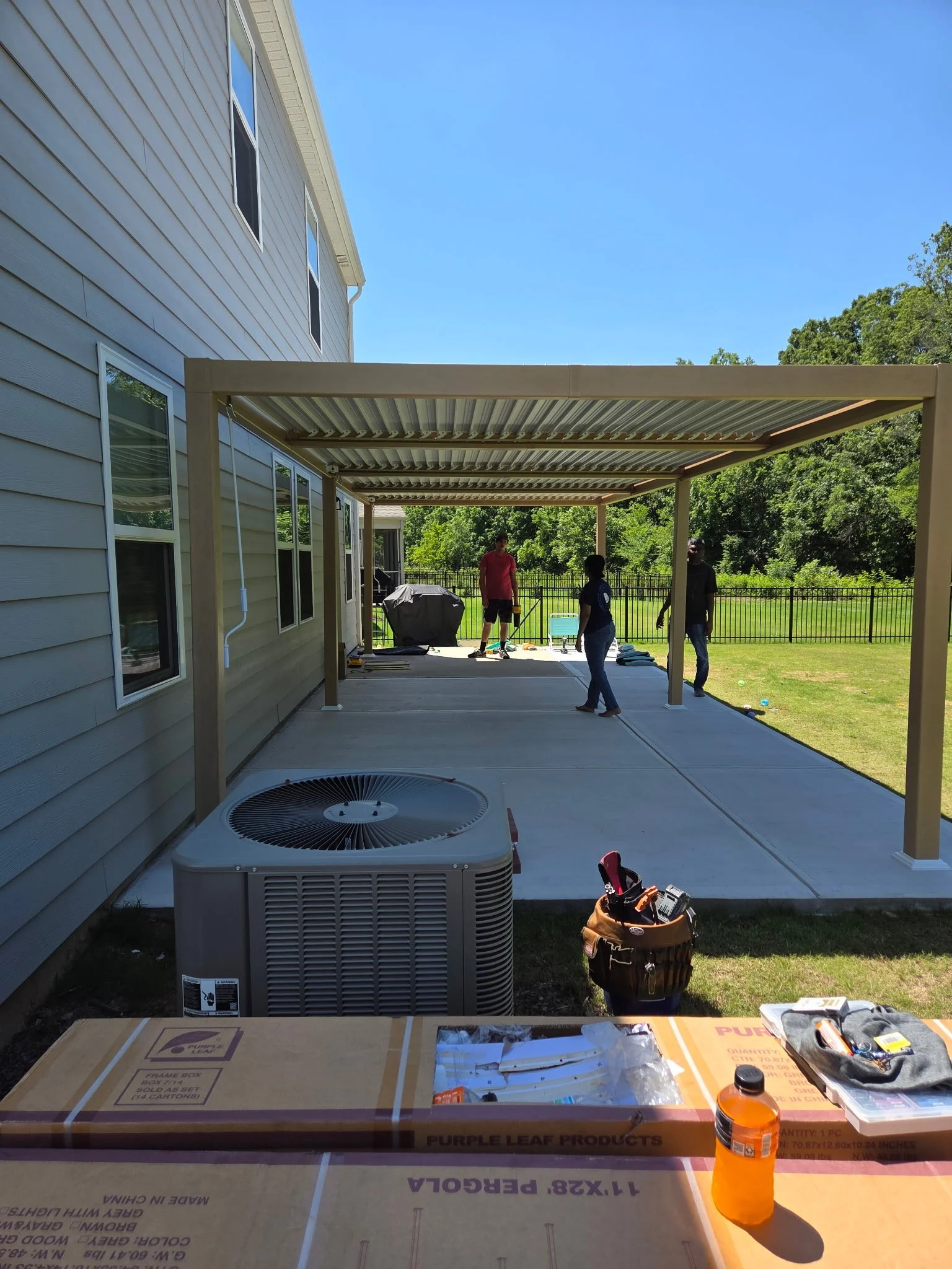 View of a backyard patio under construction, with a new metal-framed covered structure, three people working, tools and supplies scattered around, and the house's exterior siding visible on the left. There are trees and a fence in the background.