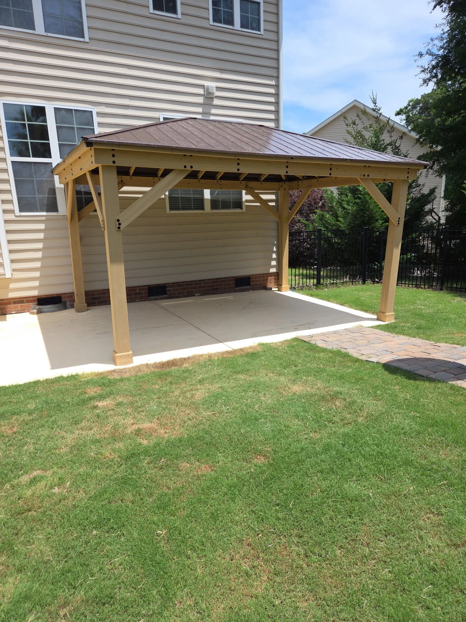 A backyard patio area with a concrete floor, a wooden pergola with a metal roof, and a grassy lawn bordered by a black metal fence. There are trees and neighboring houses visible in the background.
