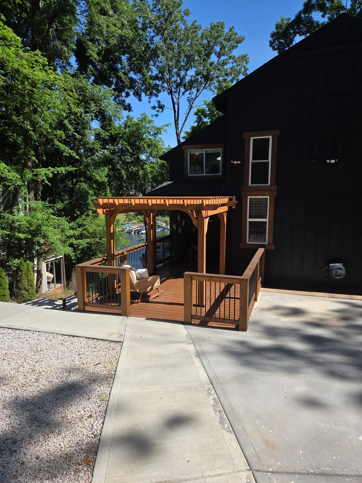 View of a house with a wooden deck and outdoor seating, surrounded by green trees, with a clear blue sky in the background.