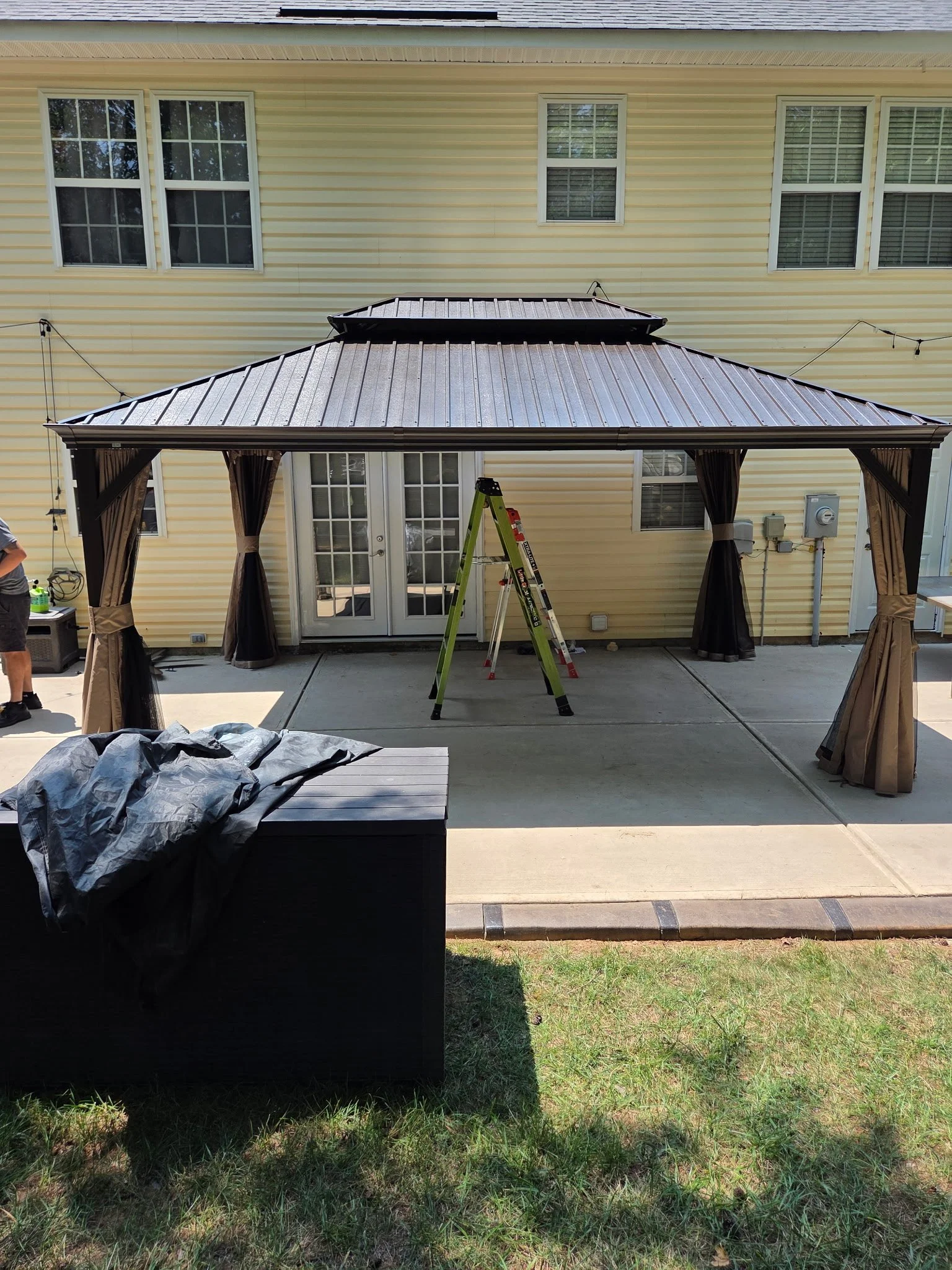 Backyard patio with a yellow house in the background. There is a metal gazebo with curtains, a ladder inside, and a black structure in the foreground. Grass and concrete ground are visible.