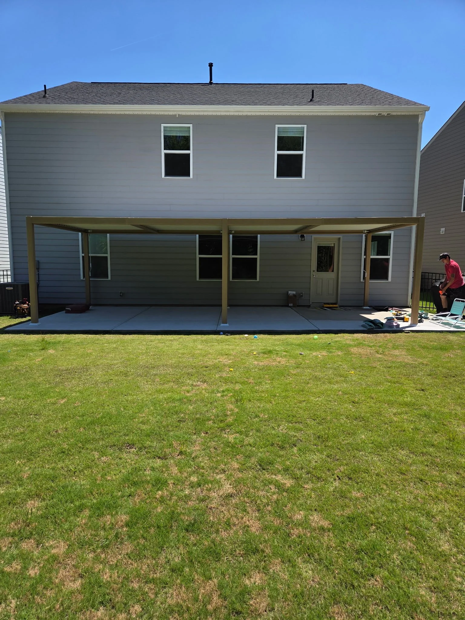 The back of a two-story house with gray siding, four windows, and a wooden patio cover. A man in a red shirt and black cap is on the right side, working on tools near a black fence. The yard has green grass and a clear blue sky.