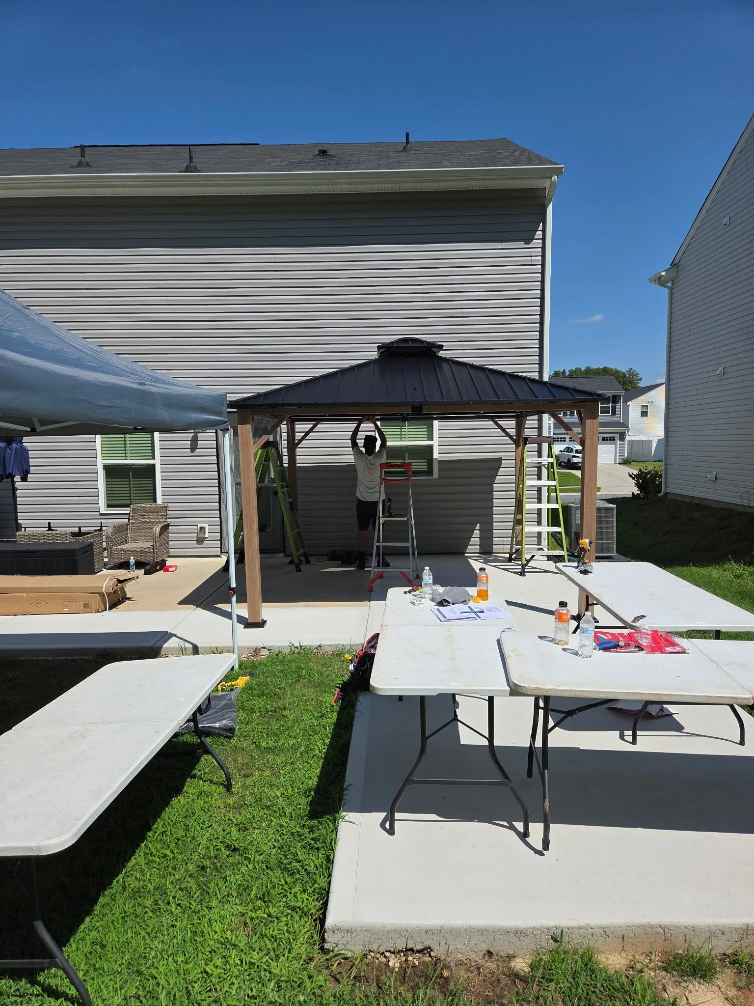 A person working on a gazebo structure outside of a house, with ladders, tools, and water bottles on tables and ground.