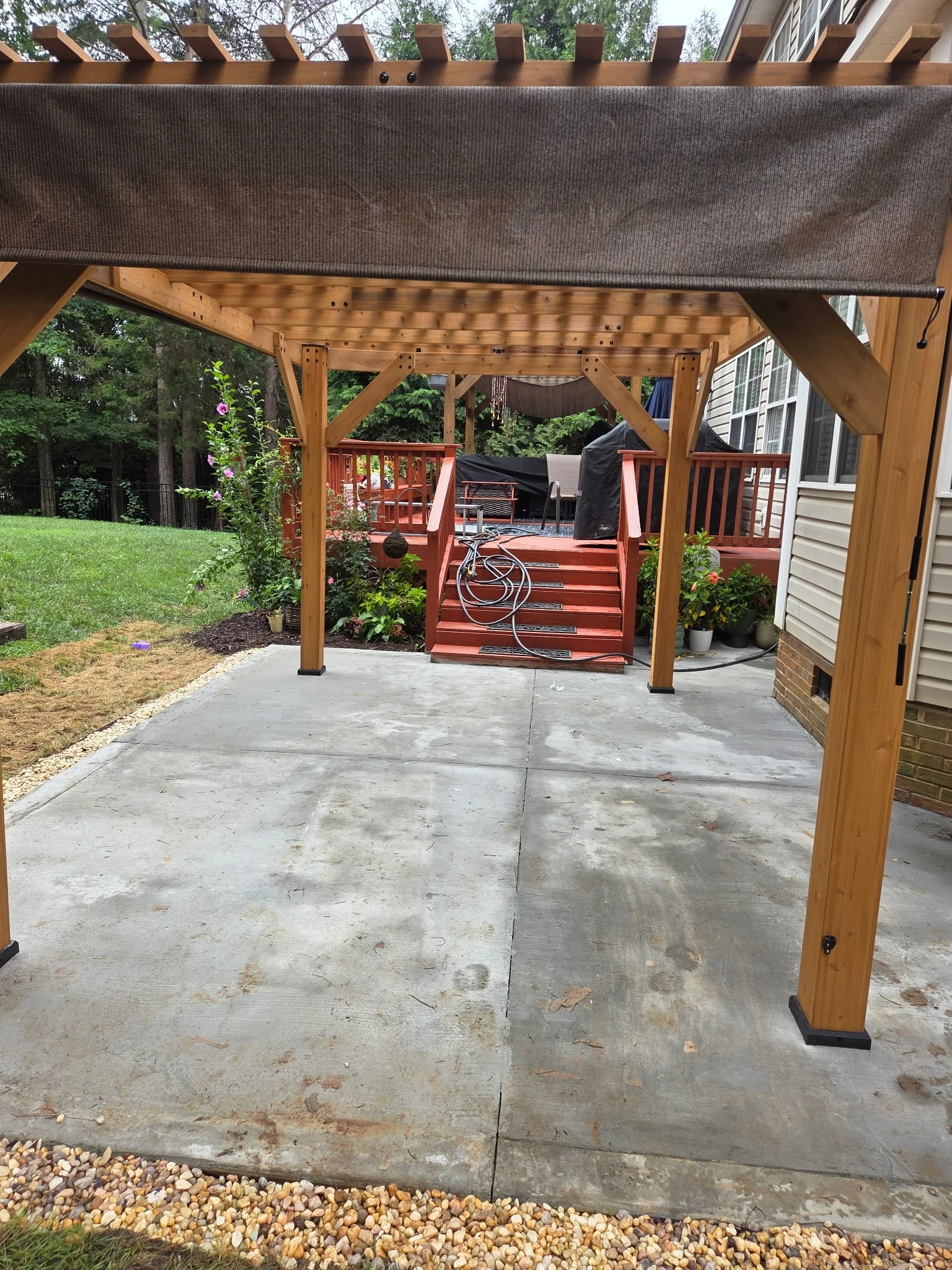 View of a backyard patio with a concrete floor, wooden deck, and a covered pergola structure. The surrounding area includes grass, plants, and trees.