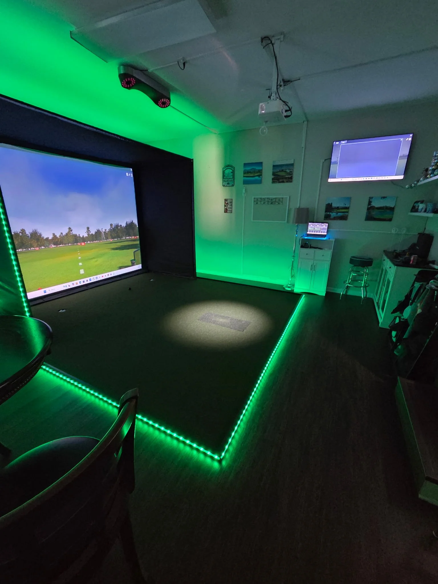 A room with green LED lighting along the edges and ceiling, featuring large display screens showing landscape images, a small table with a laptop, and chairs, creating a gaming or entertainment space.