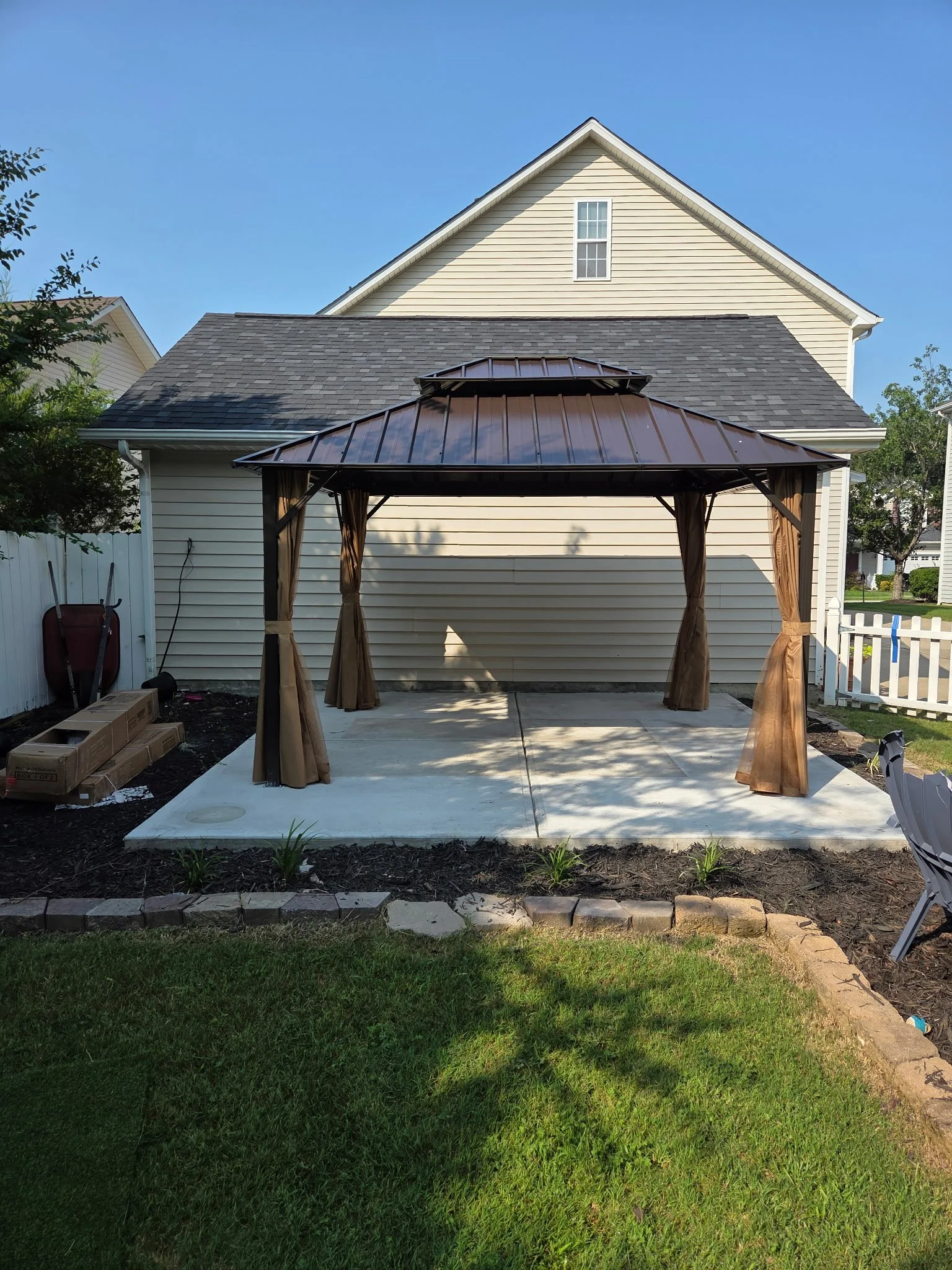 Backyard patio with concrete floor, a brown metal gazebo with curtains, small plants in the garden bed, and a house with cream siding and a pitched roof in the background.